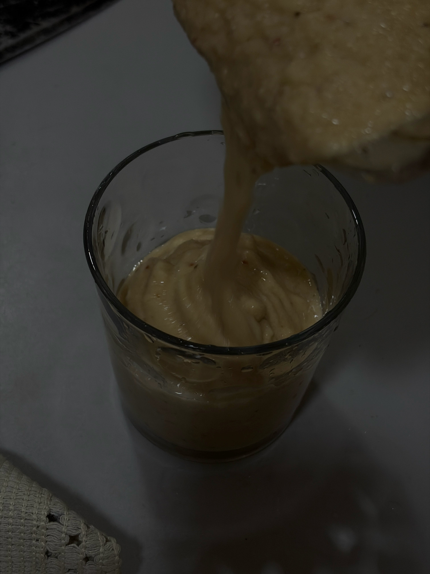 In Igboefon, Nigeria, a glass is being filled with a thick, beige-colored smoothie or milkshake from a blender. The glass is clear and shows some condensation on the exterior. The smoothie has a creamy texture with small flecks of red and brown visible. A patterned white cloth or napkin is partially visible in the lower left corner of the image. The lighting is dim, casting shadows on the surface where the glass is placed, which appears to be a countertop.