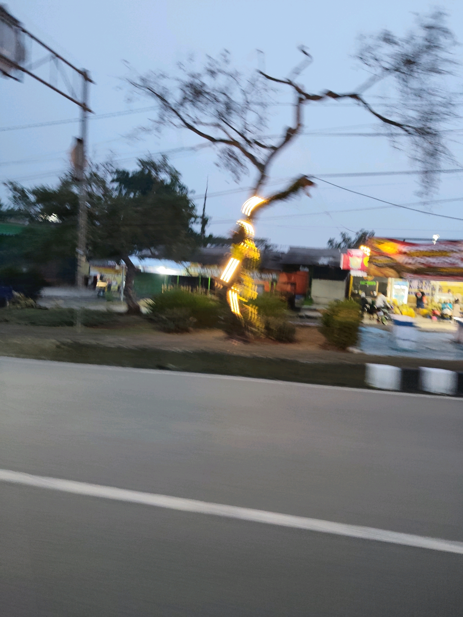 The image captures a roadside scene in Kaduagung Tengah, Indonesia. The foreground features a section of an asphalt road with a white line marking a lane. In the mid-ground, a tree adorned with yellow lights stands by the side of the road, with its bare branches silhouetted against the twilight sky. Behind the tree, a collection of small buildings or stalls are visible, some illuminated by lights. One stall appears to have a banner with text, and people can be seen gathered around. To the right, a brightly lit stall with red and yellow accents suggests a commercial area. The overall impression is a slightly blurred, motion-affected snapshot of an evening street view, likely a commercial strip or market area, common in Indonesian towns.
