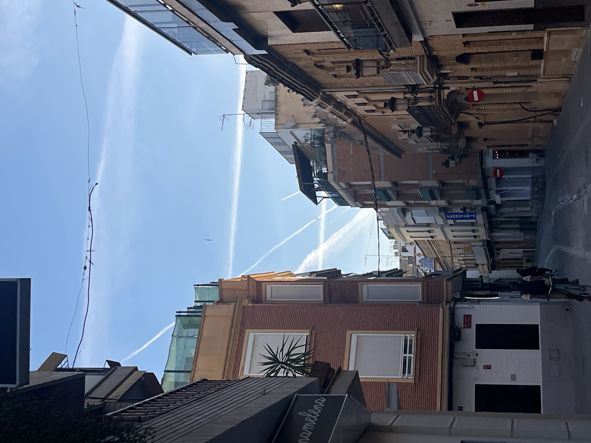 The image is of a street in Paiporta, Spain. The buildings are mostly brick and stone. There are several contrails in the blue sky. There are people on the right side of the street. A palm tree is visible on a rooftop.