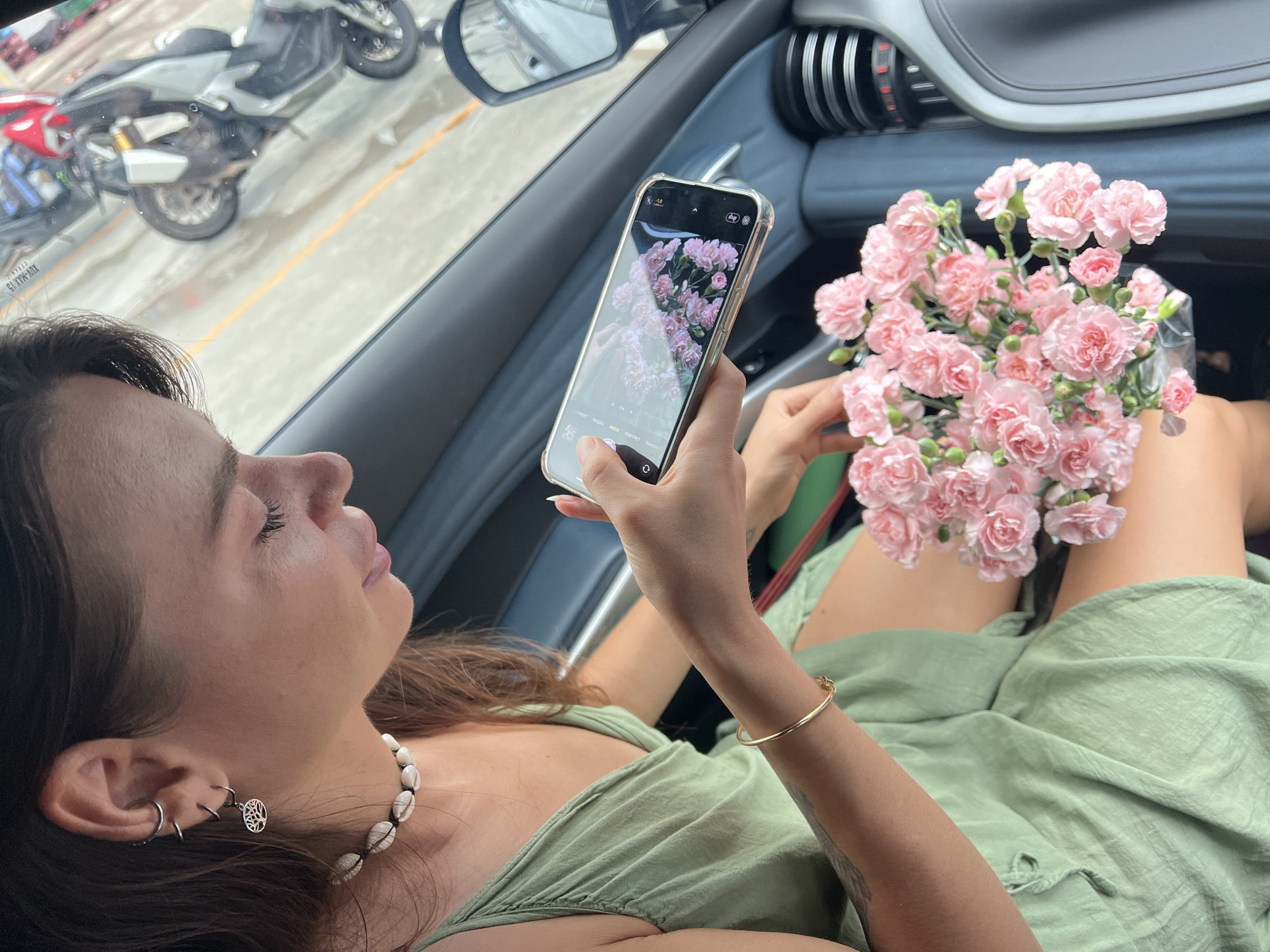 The photograph depicts a woman sitting in a car, holding a bouquet of pink flowers and taking a picture of the flowers with her smartphone. The woman is wearing a light green dress, a seashell necklace, and a gold bracelet. She has pierced ears with multiple earrings. The interior of the car is blue and silver. The background visible through the car window suggests an urban environment with parked motorcycles. Based on the location in the prompt, Baan Chaweng, Thailand, it's likely a street scene in that area.