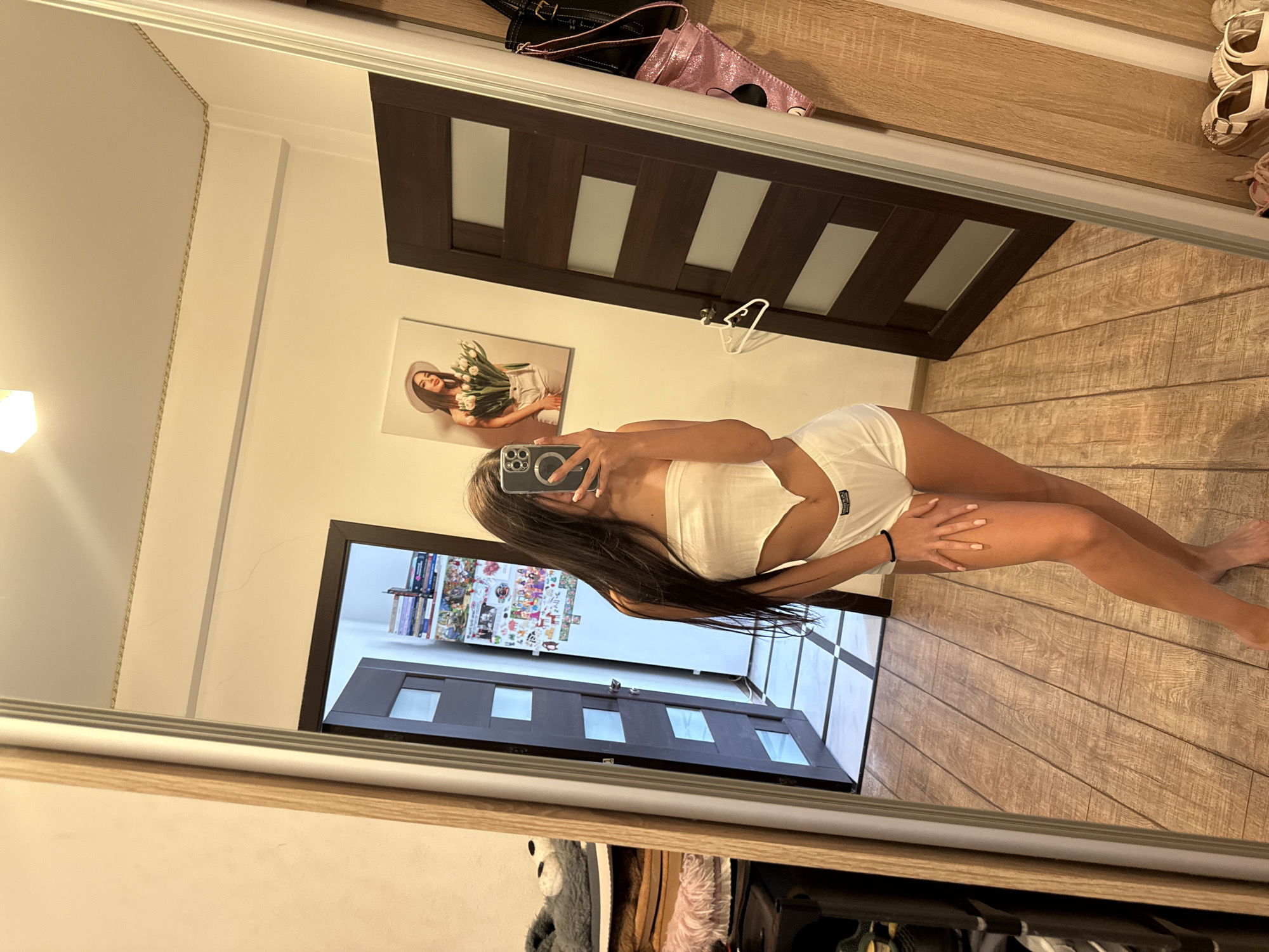 A young woman is reflected in a mirror, wearing a white bandeau top and white shorts. She is holding a smartphone in her right hand, using it to take a selfie. The background shows a room with wooden floors and dark brown doors. One door has a picture hanging on it of a woman holding tulips. The other door is open, revealing a tiled hallway. To the left of the woman, there are shelves with various items, including plush slippers and folded blankets. The overall impression is a candid snapshot taken in a private space, likely a bedroom or dressing area. The image does not provide any specific geographical markers that would indicate the location in Chișinău, Moldova.