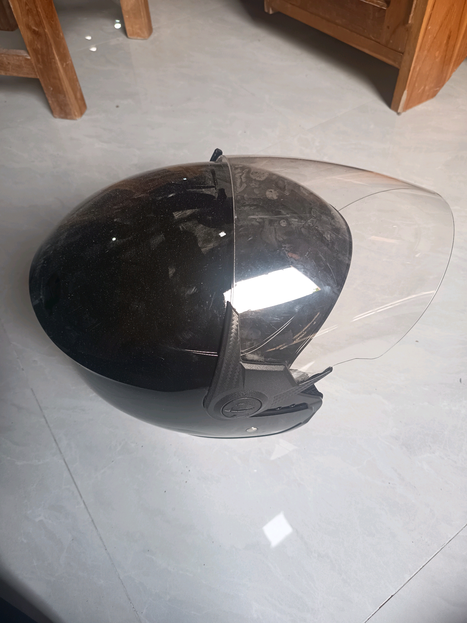A black helmet with a clear visor rests on a tiled floor. The helmet is dusty and appears to be a half-helmet style with a carbon fiber accent near the visor hinge. The floor is light gray with a subtle marble pattern. In the background, wooden furniture, possibly a cabinet and chair legs, are visible. The scene suggests an indoor setting. The overall impression is of an object placed on the floor in a domestic or storage area. The location is Sidorejo, Indonesia.
