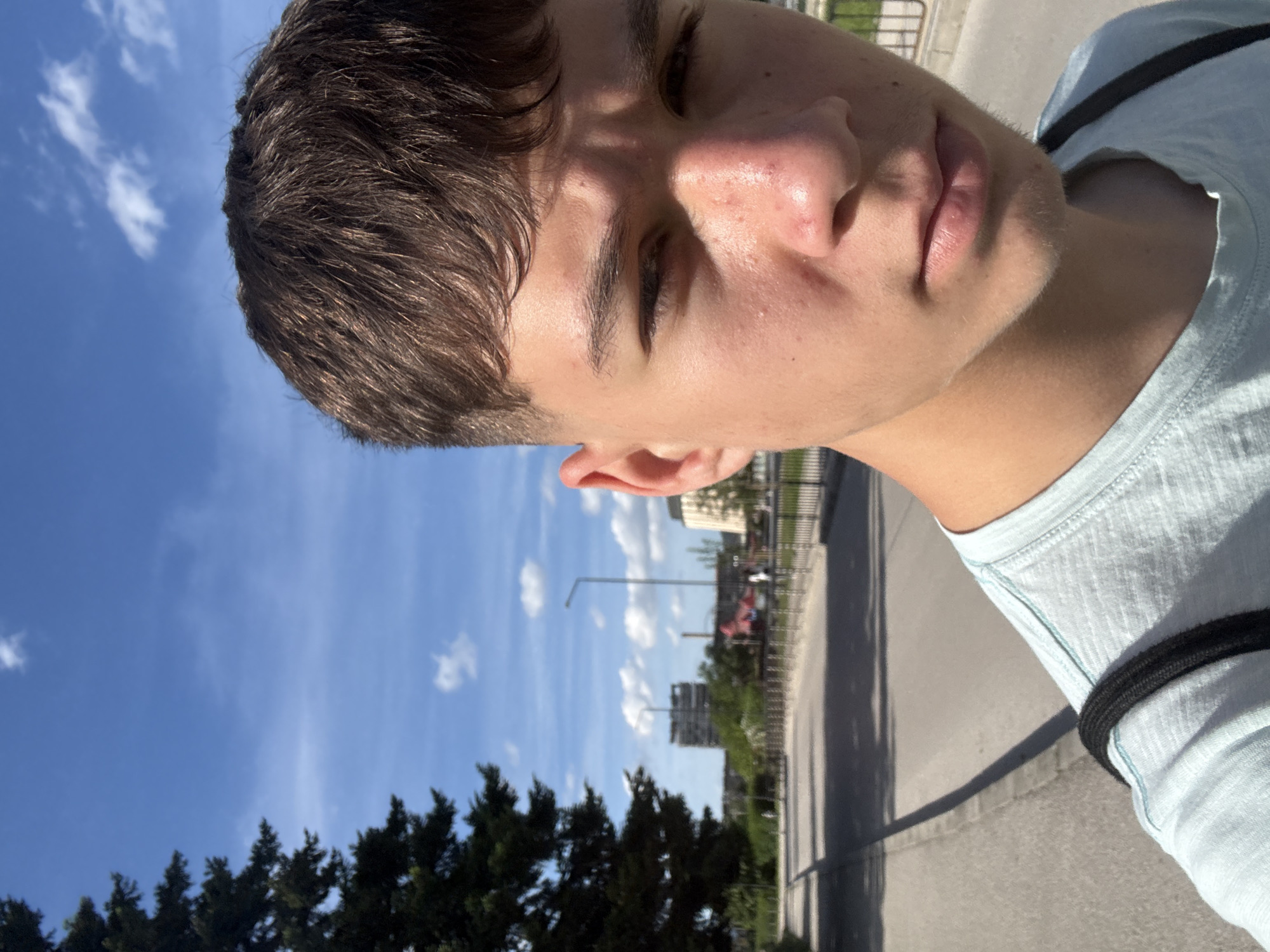 A close-up, tilted selfie of a young man with short, dark brown hair and fair skin. He wears a light blue t-shirt and is outdoors. The background shows a blue sky with scattered clouds, tall green trees, a gray road or sidewalk, and some buildings, including what appears to be a light pole and other urban structures. The exact location within Gabrovo, Bulgaria, cannot be determined with the available information.