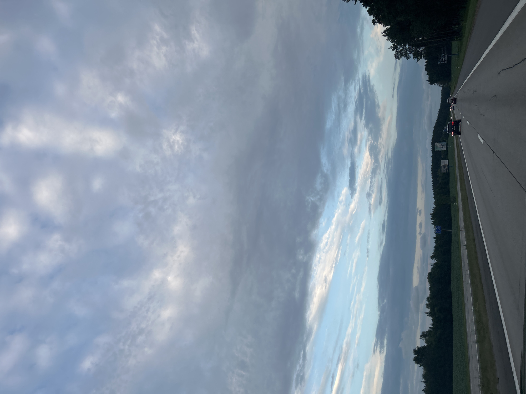The image depicts a highway scene in the Ozyaritska-Slabadski rural council, Belarus. The sky is filled with a mix of light and dark gray clouds, with patches of blue sky visible. The highway has at least two lanes in each direction and is bordered by grass and trees on both sides. On the left side of the highway, there are fields and a dense forest. On the right side, there is a thick forest. Several cars are traveling on the highway, including a dark SUV in the foreground. There are also some road signs visible, including a blue sign with the number "13" on the left side of the road. The overall impression is a travel scene on a cloudy day in a rural area.