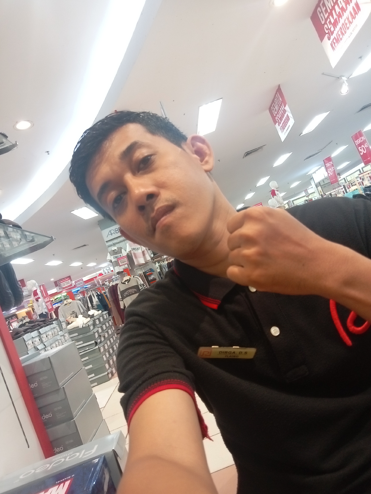 In a retail environment, likely a department store, a male individual with dark hair and brown eyes takes a selfie. He is wearing a black polo shirt with red trim and a name tag that reads "DIRGA. D.S." The store appears to be decorated with red and white banners, some of which contain text in Indonesian, suggesting a celebratory event. The background reveals racks of clothing, stacks of shoeboxes, and fluorescent lighting common in such establishments. The presence of Indonesian text on the banners and the name tag suggests the location is in Indonesia, potentially in the city of Batam, as indicated by contextual information. The overall scene is well-lit and shows a consumer-oriented setting.