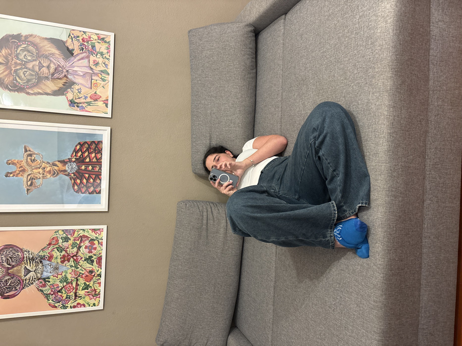 An indoor scene in Nesso, Italy, depicts a young adult female reclining on her left side on a grey fabric sectional sofa. She is dressed in a white short-sleeved t-shirt, dark blue wide-leg denim jeans, and a single bright blue slipper with white detailing on her visible right foot. Both hands grasp a grey smartphone, equipped with a white circular magnetic accessory, and her gaze is directed at the screen. Above the sofa, mounted on a plain light brown wall, are three framed art prints featuring anthropomorphic animals wearing suits and eyeglasses: from left to right, a leopard in a floral suit, a giraffe in a patterned suit, and a lion in a floral suit with a pink cravat. The room is uniformly lit.