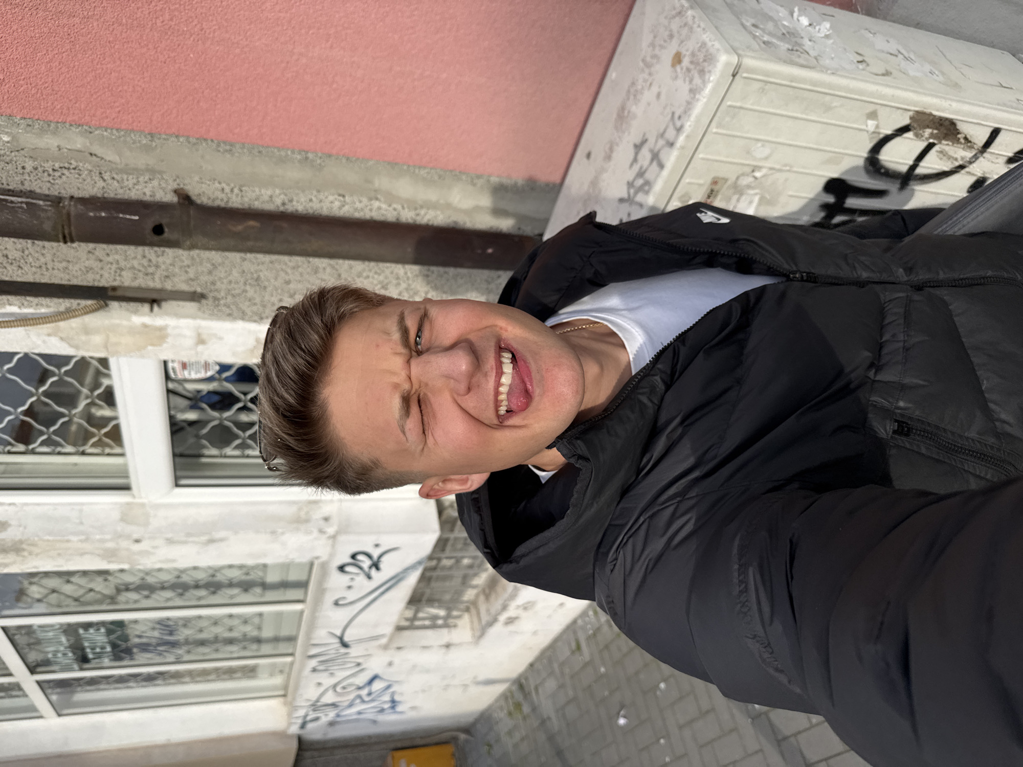 A selfie taken at street level in Burgas, Bulgaria shows a young man with light brown hair wearing a black puffer jacket and white t-shirt. He is making a silly face, squinting his eyes, sticking his tongue out, and smiling. The background includes a building with a salmon-colored wall and a white building section with graffiti. A window with decorative metal grilles is visible. The sidewalk is paved with gray tiles. There's a drainpipe along the salmon-colored wall and electrical conduit.