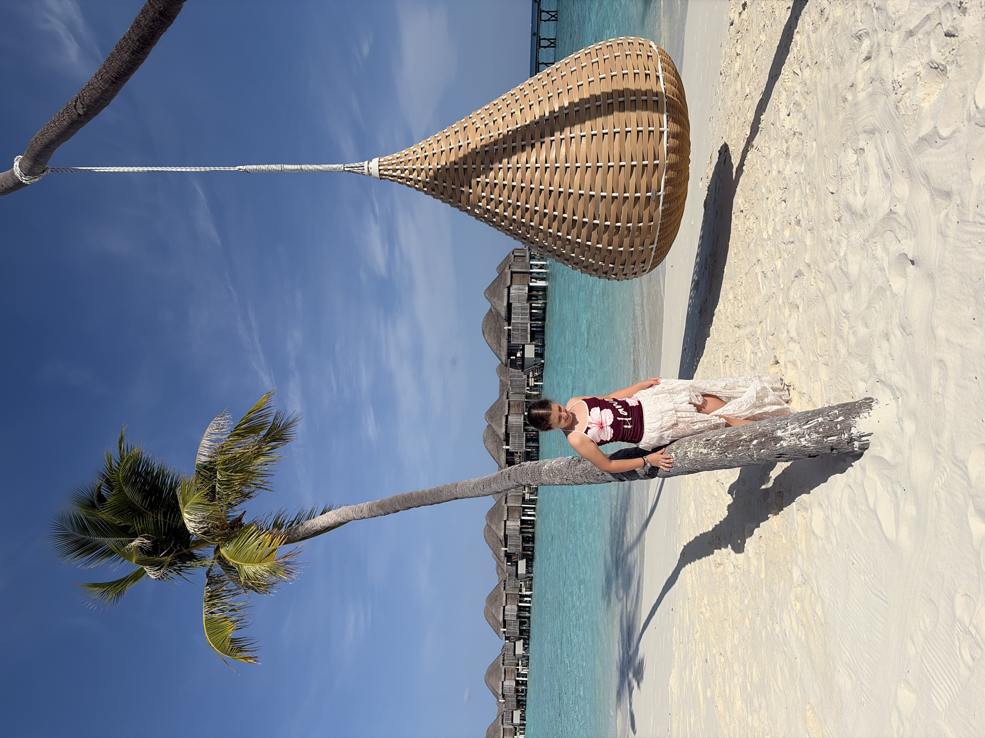 A young woman stands leaning against a palm tree on a sandy beach. She is wearing a dark red tank top with a floral design and the word "Hawaii" and a flowing, light-colored skirt. To her right, a large, teardrop-shaped woven swing hangs from a rope attached to a tree branch. In the background, turquoise water separates the beach from a row of thatched-roof overwater bungalows. The sky is a clear, bright blue with a few wispy clouds. The sun casts long shadows from the palm tree onto the sand. The scene is one of a tropical paradise in Halaveli, Maldives.