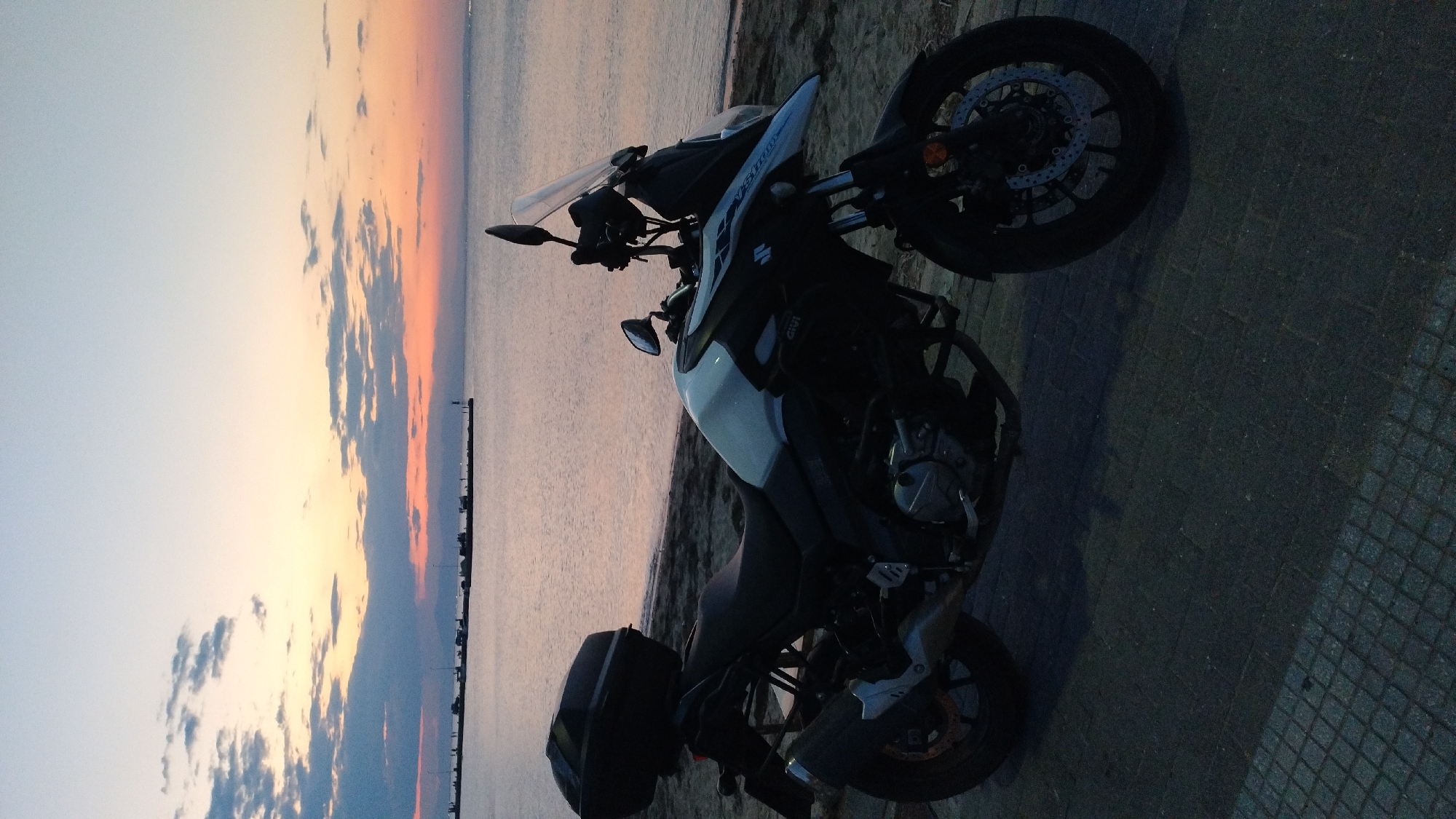A black and silver Suzuki V-Strom motorcycle with a black top case is parked on a paved walkway next to a sandy beach. The motorcycle is positioned in the foreground, facing right. Behind the motorcycle, a calm body of water stretches to the horizon. The sky above is a gradient of pale blue and orange, with scattered clouds, indicative of either sunrise or sunset. In the distance, a pier with silhouetted figures and structures extends into the water. The setting appears to be a coastal area, potentially the promenade in Neoi Epivates, Greece, given the presence of a beach, water, and a pier. No people are actively engaged in any actions. The environment is peaceful and still.