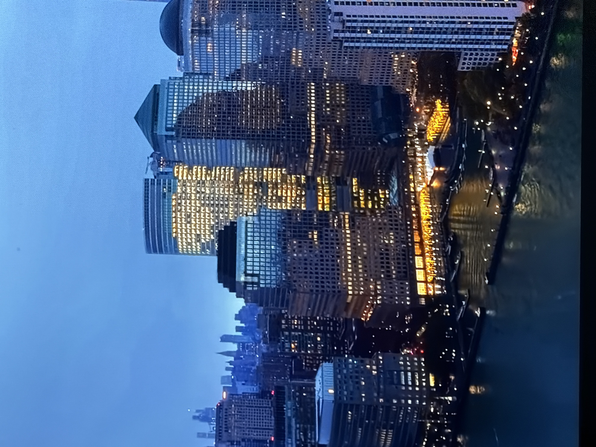 A detailed aerial view captures a densely developed urban waterfront area in the city of Maidstone, United Kingdom, at twilight or night. The scene is dominated by a cluster of illuminated skyscrapers and commercial buildings lining a wide body of water, under a deep blue sky.
In the midground, numerous multi-story buildings stand prominently. Several central structures feature distinct architectural tops, including dark pyramidal and light blue domed roofs. One large building on the left is almost entirely illuminated with warm yellow lights, creating a grid pattern visible through its windows. Other buildings display a mix of lit and unlit windows, reflecting the blue sky. In the far background, a hazy outline of additional, smaller skyscrapers is visible against the horizon.
The lower portion of the image shows a wide body of dark blue-green water, reflecting the bright lights of the city above. Along the waterfront, a promenade is delineated by numerous small, bright artificial lights. Several dark, elongated structures, appearing to be piers or docks, extend into the water, with what seem to be small boats or barges moored alongside them. Buildings fronting the water exhibit illuminated ground-level windows, suggesting commercial or retail spaces. The sky above is a clear, deep blue, with a faint, small dark spot present in the upper central area. No specific individuals or actions are discernible in the scene.