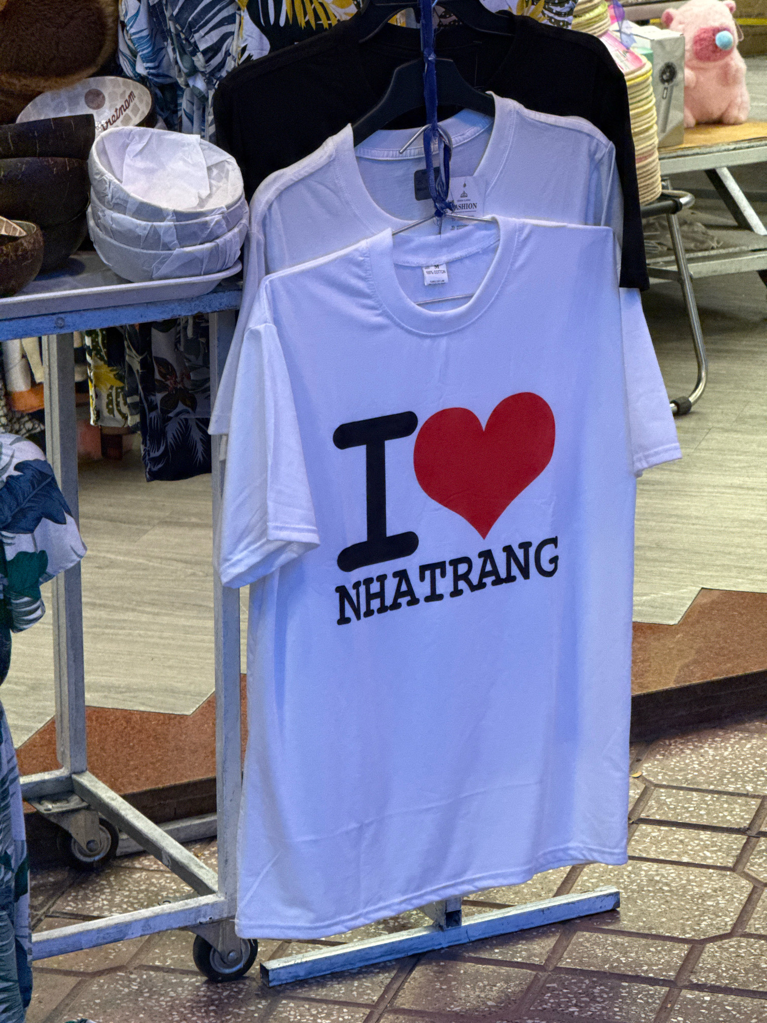 This image shows a close-up view of a white t-shirt with the text "I ❤️ NHATRANG" printed on it, hanging on a clothing rack. The t-shirt is the primary subject. The background includes other clothing items, a pink stuffed animal, and a tiled floor. The scene appears to be a market stall or shop in Nha Trang, Vietnam. The overall lighting suggests it's daytime, possibly indoors or under shade.