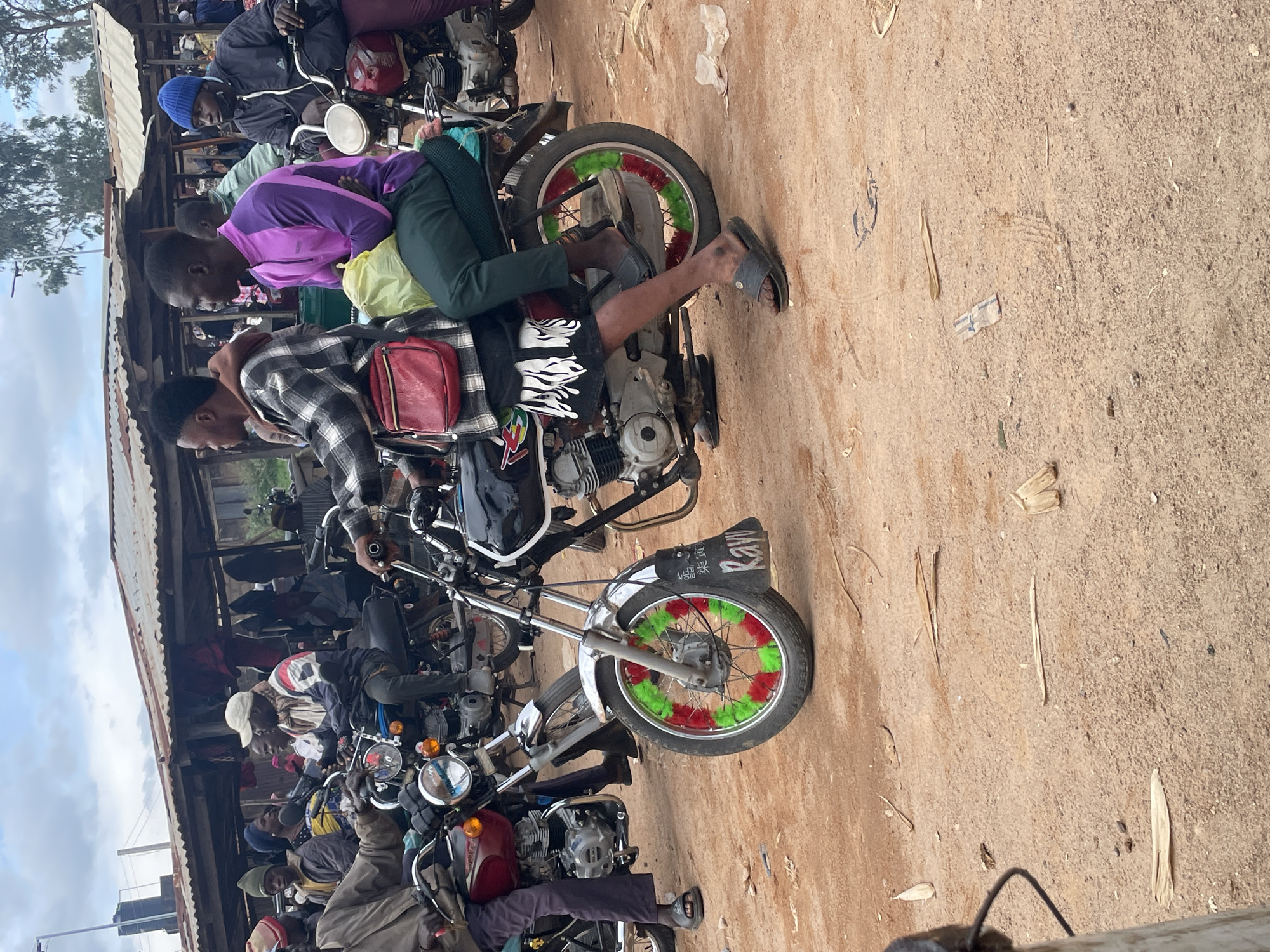 The image depicts a street scene in Vom, Nigeria, likely a market or roadside area. In the foreground, two men are seated on a motorcycle. The driver, in a black and white checkered shirt, has a red bag slung across his chest. The passenger, in a purple and pink long-sleeved shirt, is holding a yellow plastic bag. Both men are wearing casual footwear. The motorcycle has a fender decorated with green and red pom-poms and a sign with the word "Raw" written on it.
Behind them, several other individuals are visible, some on motorcycles, in a busy, unpaved environment. Several stalls with corrugated metal roofs are present in the background, suggesting a commercial area. The ground is dusty, with scattered dry leaves and debris. The overall impression is one of daily life and commerce in a Nigerian town.