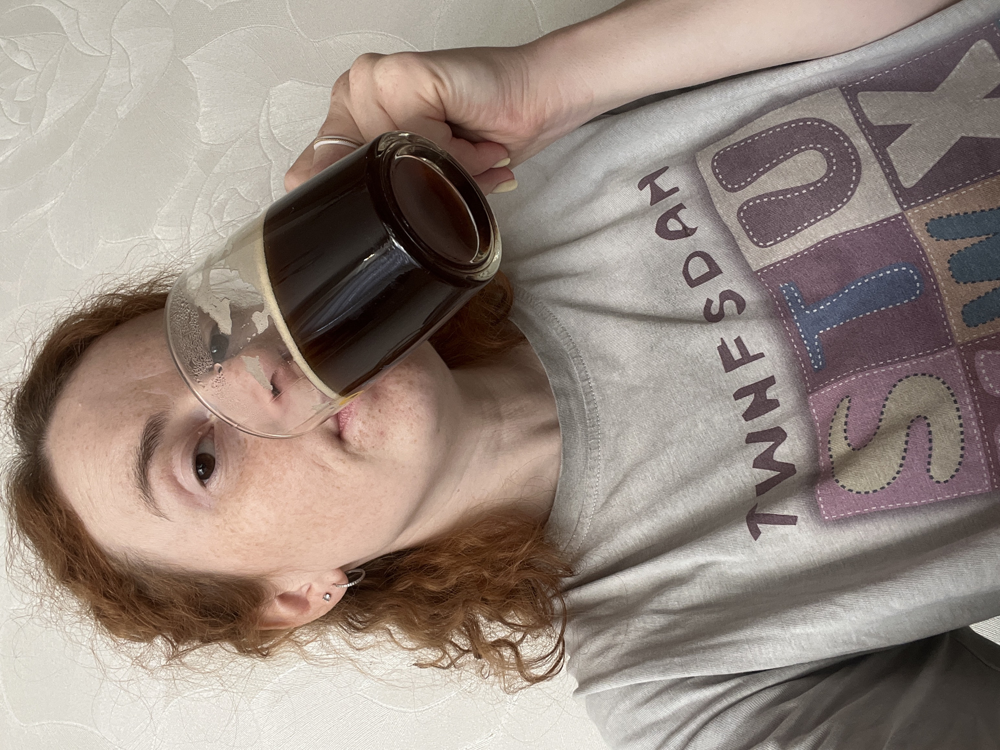 A person with red hair is drinking from a clear glass filled with a dark liquid, likely coffee. They are wearing a gray t-shirt with the word "Thursday" printed vertically down the front. The person is fair-skinned with freckles. They have a ring on their finger and an earring in their ear. The background is a textured white surface, possibly a wall or bedding. There is no information to indicate the location of the image to be in the city of Chaikovsky, Russia.