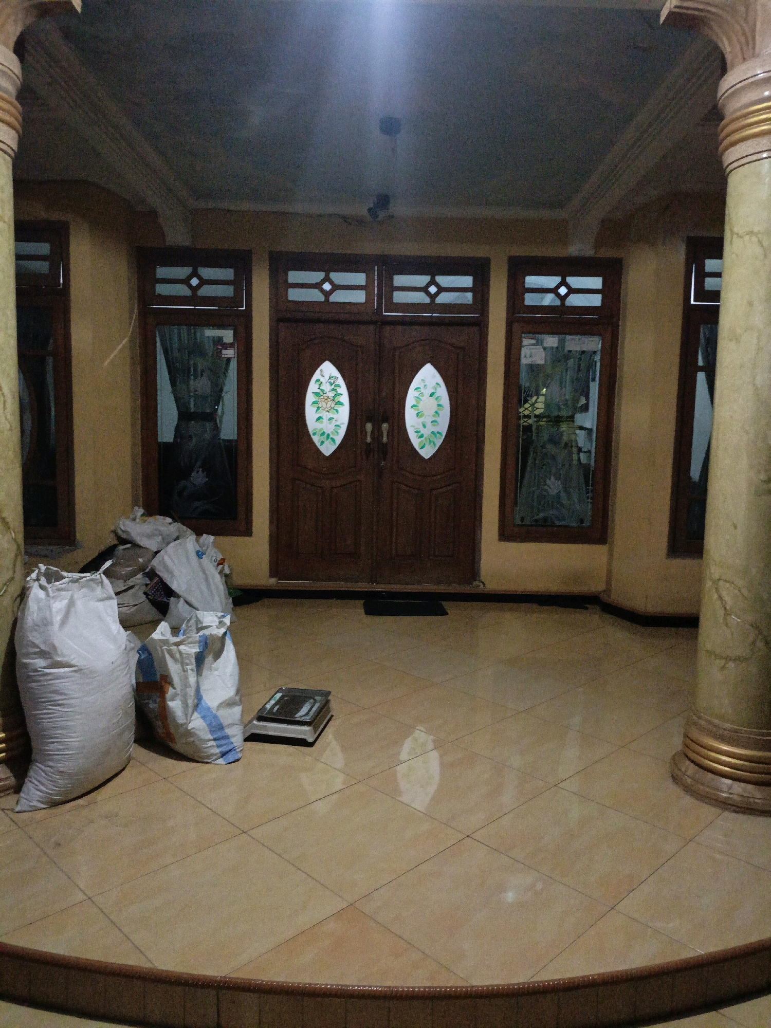 The image shows the entrance area of a building, likely a residence, in Betek, Indonesia. The scene is dimly lit, suggesting it might be evening or indoors with artificial lighting.
Dominating the center of the image are a pair of large, wooden double doors, ornately carved and featuring decorative oval inlays with floral designs. To the left and right of the doors are tall, narrow windows with wooden frames and patterned glass or curtains, reflecting some light.
The floor is tiled with a light brown, glossy material, showing reflections of the lighting and the environment. On the left side of the frame, near the entrance, are several large, white woven sacks, some appearing to be partially filled. Beside these sacks, on the floor, is a small, dark-colored weighing scale.
The entrance is flanked by two large, decorative pillars. The pillar on the right is particularly visible, with a textured, marble-like finish and a golden, tiered base. The pillar on the left is partially obscured by the sacks. The ceiling above the entrance has exposed beams and appears to be a darker color, possibly concrete.
The overall impression is of a traditional Indonesian home entrance, with a mix of utilitarian items like the sacks and scale, and decorative elements like the doors and pillars.