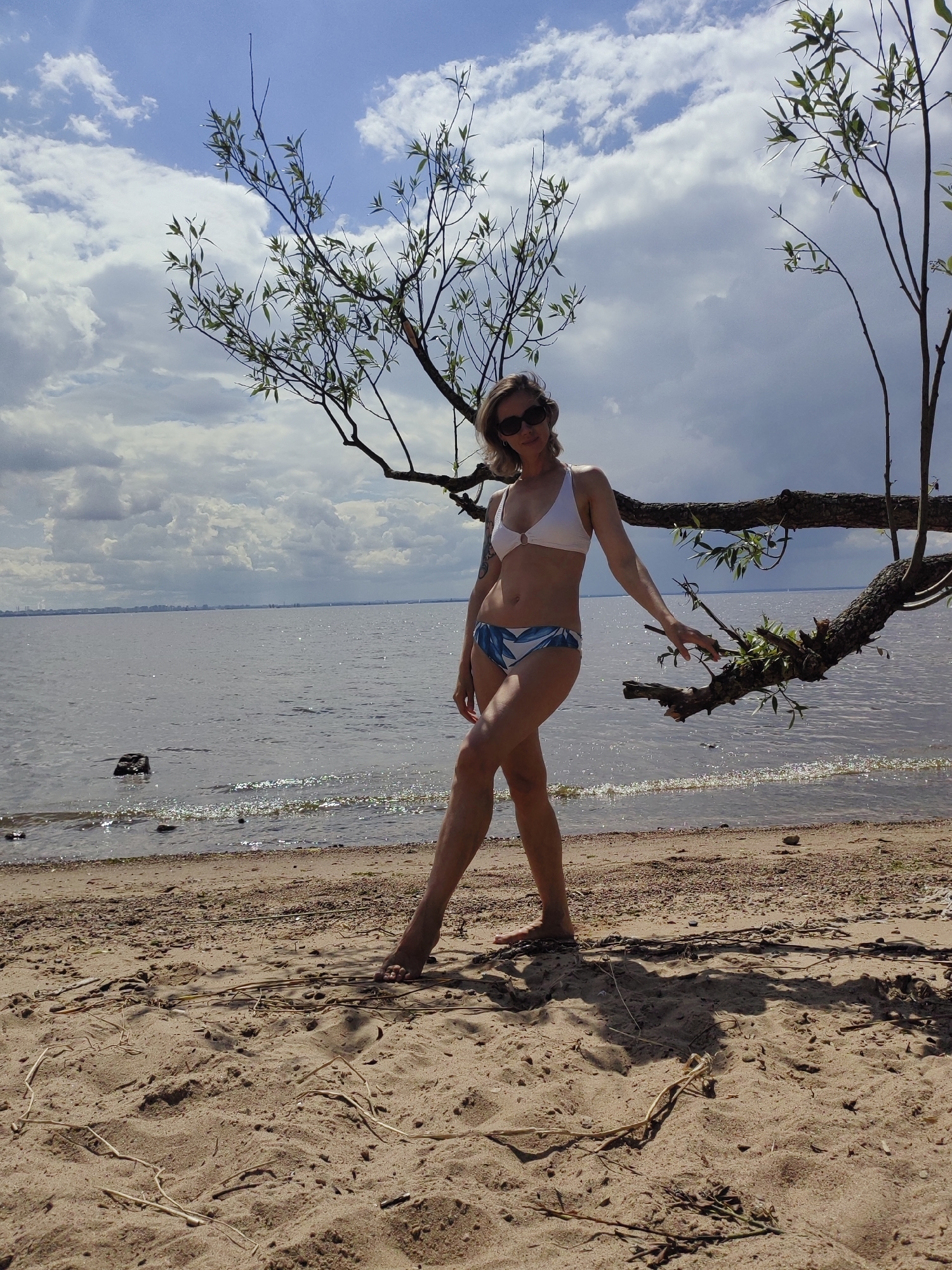 A woman in a white bikini poses on a sandy beach beside a body of water. She has blonde hair, sunglasses, and is wearing a white top with a ring detail and blue and white patterned bottoms. A tree branch with green leaves extends from the right side of the frame, hanging over the water and the woman. The water is calm with a slight shimmer from sunlight. The sky is partly cloudy with white and grey clouds. The beach is sandy with some scattered pebbles and dried plant material. The scene appears to be outdoors on a sunny day. Based on the landscape visible in the background, this location is likely the Gulf of Finland coast near Saint Petersburg, Russia, possibly near Sestroretsk or Komarovo, known for its beaches and pine forests.