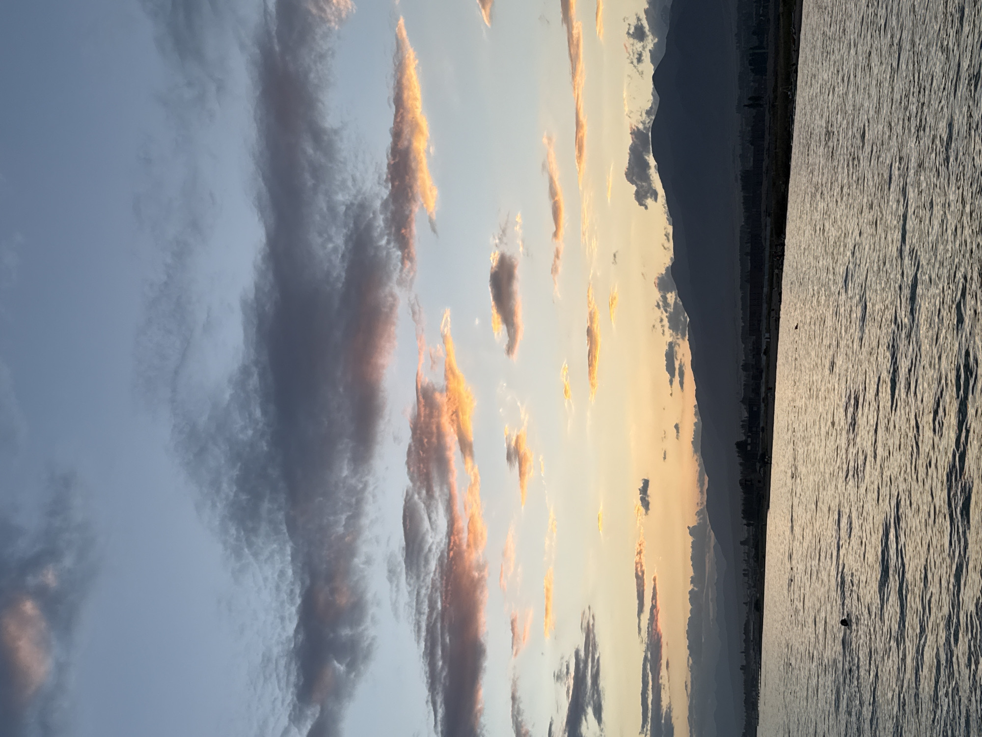 The image captures a serene sunset over the Issyk-Kul lake, near the village of Sary-Oy, Kyrgyzstan. The sky is a canvas of soft blues and oranges, with streaks of pink and gold illuminating the scattered clouds. Below, the tranquil waters of the lake reflect the warm hues of the sky, creating a shimmering surface. A lone swimmer is visible in the water, a small dark silhouette against the vast expanse. The distant shoreline is lined with dark, silhouetted mountains, their rugged peaks fading into the dusky light. A narrow strip of land separates the lake from the mountains, featuring scattered trees and buildings that are indistinct in the fading light. The overall atmosphere is one of peaceful solitude and natural beauty.