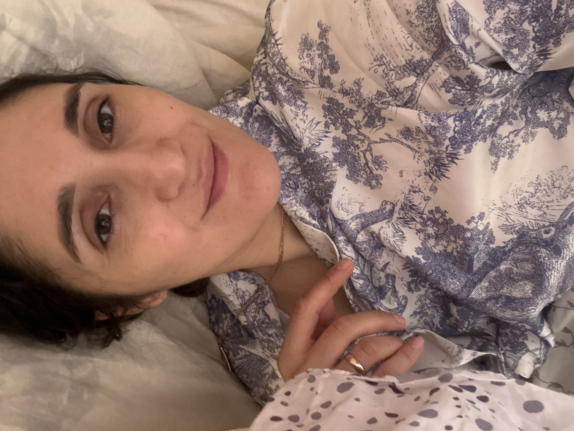 A person is lying on a bed covered with a white sheet, facing the camera. The person has brown hair and eyes. The person is wearing a patterned blue and white shirt. A gold ring is visible on their finger. A gold chain necklace is around their neck. The location is not discernible, and there is no indication of it being in Tobolsk, Russia.