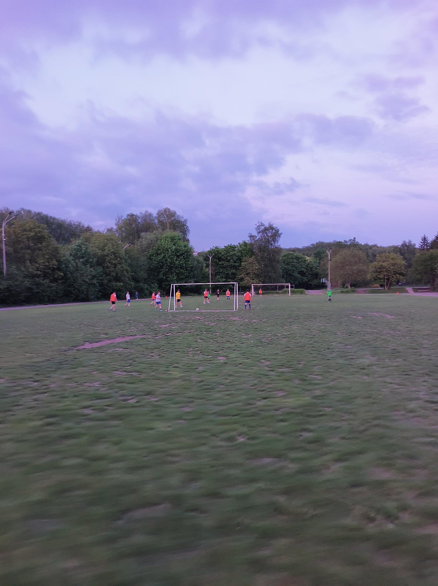 The image shows a soccer field in Ternopil, Ukraine. There are several people playing soccer, and two soccer goals are visible. The sky is cloudy, and the surrounding area is forested.
