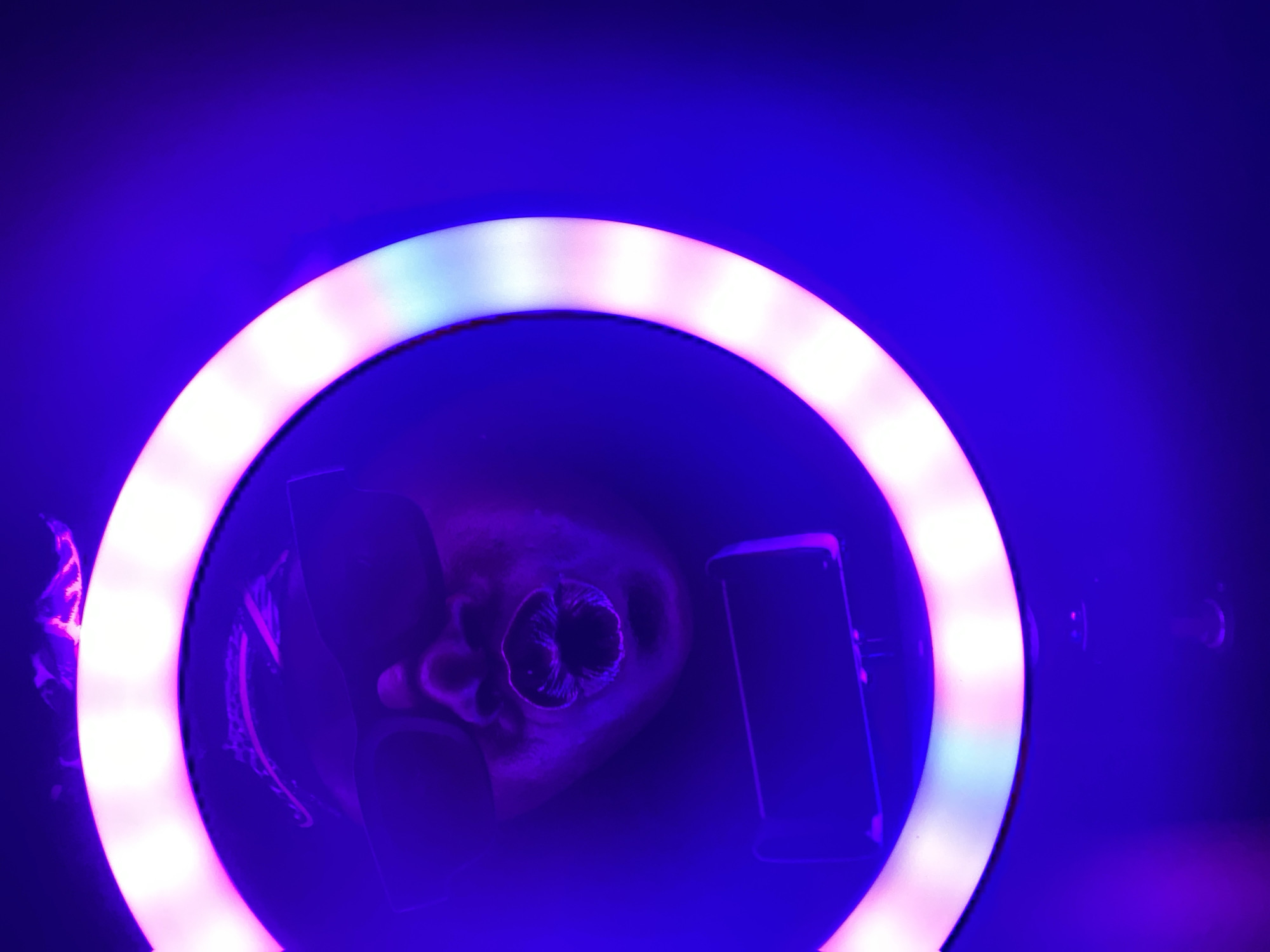 The image presents a top-down view into the center of a brightly lit ring light, bathed in an ambient deep blue and purple hue.
**Subjects:** The primary subject is a person's head, seen from above, positioned within the inner circle of the ring light. The features of the face are dimly visible, mostly illuminated by the purple light and some glow from the ring light, making specific details difficult to discern, though what appears to be the nose and mouth area are somewhat clear. A dark, rectangular object, likely a smartphone or a small camera/monitor, is positioned vertically to the right of the person's head, also within the ring light's circle.
**Activity or event taking place:** The setup strongly suggests a photographic or videographic session, possibly a selfie or a video call, where the ring light provides illumination for the person looking into the camera (presumably the device within the ring). The prominent purple lighting indicates a deliberate aesthetic choice.
**Notable details:** The most striking feature is the vibrant and pervasive blue and purple lighting that saturates the entire image. The ring light itself emits a bright white light, which creates a halo effect and distinct segments are visible along its circumference. The contrast between the bright white light of the ring and the deep blue/purple background and subject creates a somewhat surreal and high-tech atmosphere. No specific location cues are visible within the image to directly link it to Maigizo, Nigeria. The lighting suggests an indoor setting or a night-time scenario. No visible text is present in the image.