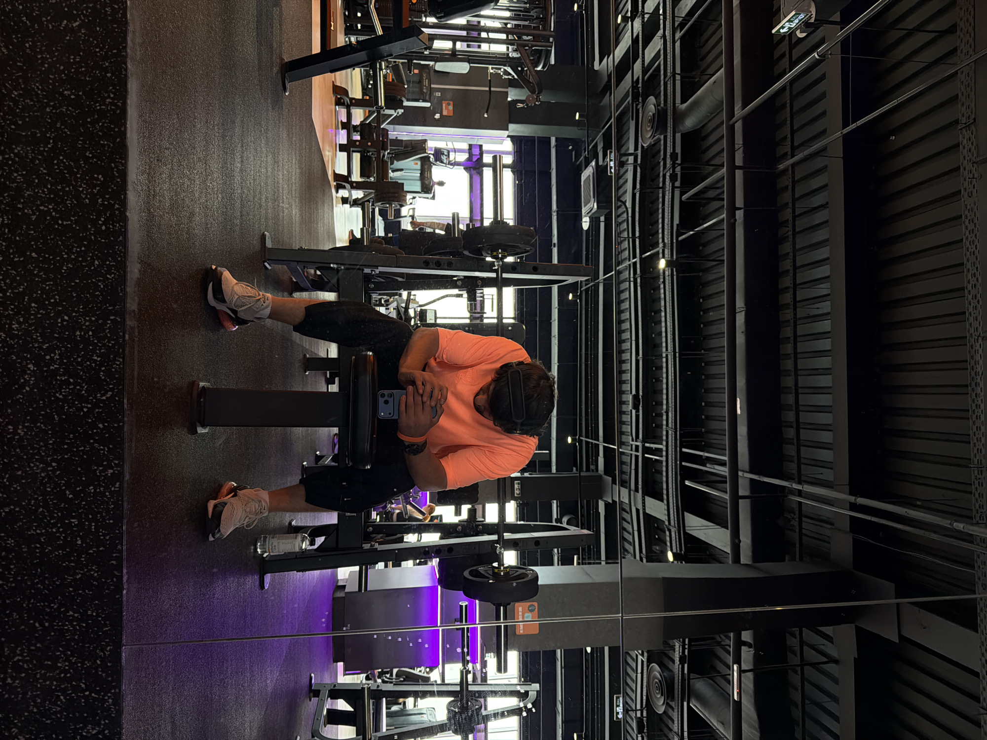 A person is sitting on an exercise machine in a gym. The individual is wearing an orange t-shirt, black shorts, and headphones, and holding a blue phone in their hands. The gym contains weightlifting equipment, with a black floor and a dark ceiling with exposed pipes and ventilation systems. The person's left foot is facing away from the machine, and their right foot is pointing towards the machine. The scene appears to be well-lit with some purple accent lighting. There is no verifiable evidence from this image to pinpoint the exact location in Krasnoyarsk, Russia.