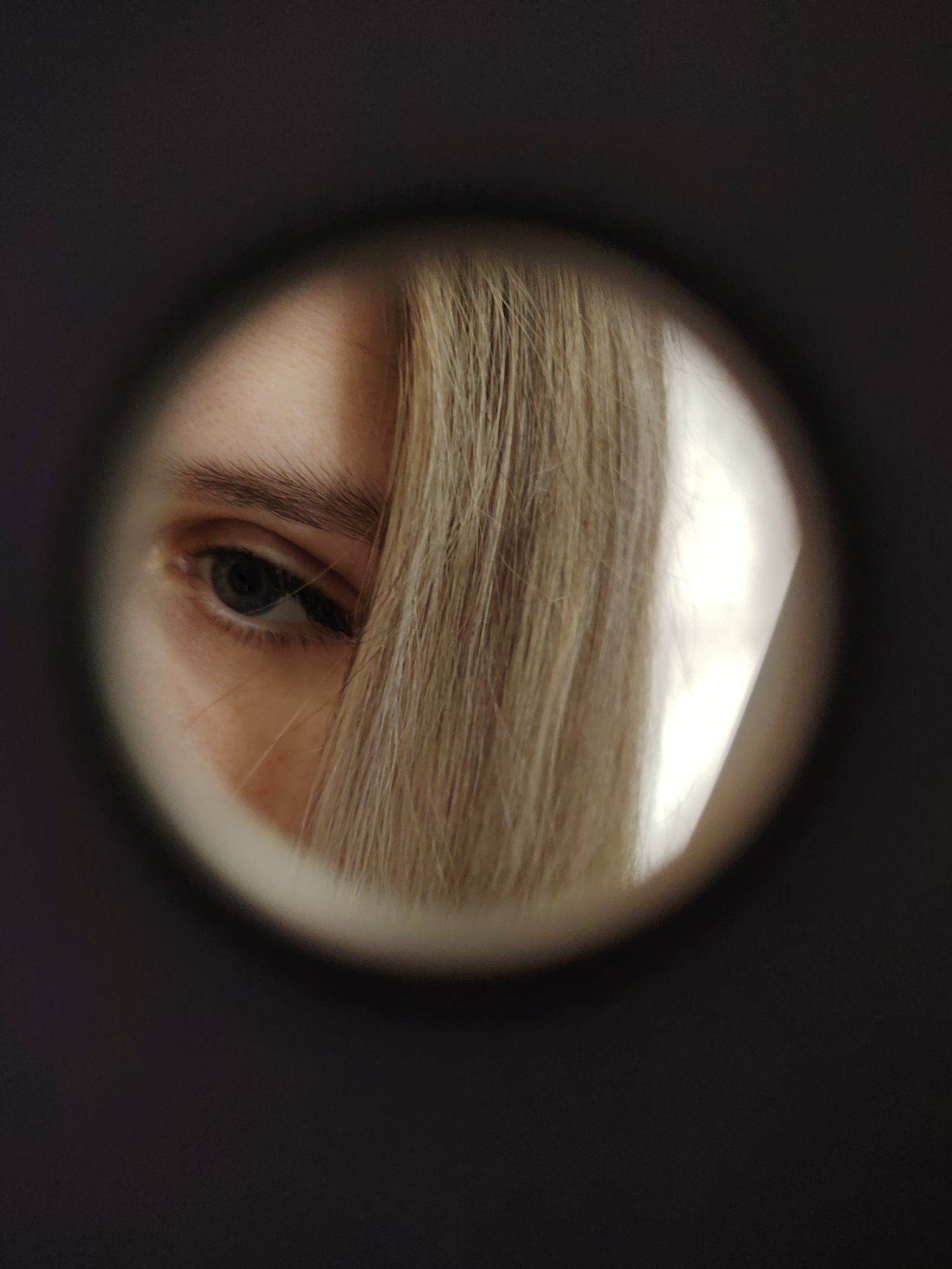 The image shows a close-up of a person's eye and hair seen through a peephole. The person has light-colored hair partially obscuring their face. The eye is visible and gazing to the left. The peephole creates a circular frame around the eye and hair, set against a dark background. The location in the city of Chaikovsky, Russia, is not discernible from the visual information provided in the image.