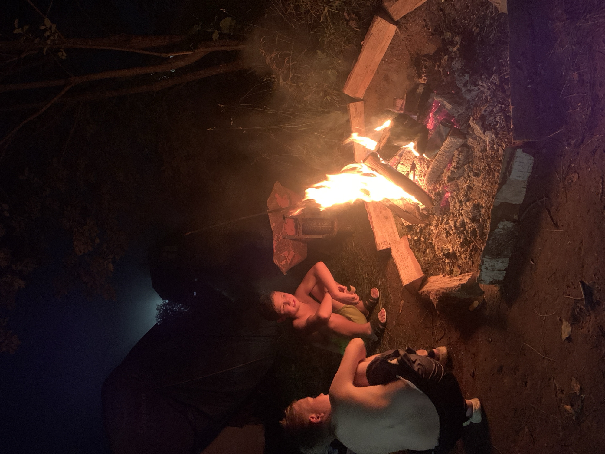 Two young boys are sitting around a campfire at night. The boy on the left is facing away from the camera, looking towards the fire. The boy on the right is facing the fire and looking slightly to his right. They are both shirtless and wearing shorts. The campfire is burning brightly, with flames reaching upwards. There are several logs placed around the fire. In the background, a tent is visible to the left, and trees are present to the right. The ground is covered in dirt and grass. The sky above is dark. This scene appears to be in an outdoor setting, possibly a campsite. It is dark, suggesting it is evening or night. There is no specific indication of the location within Zasechnyi, Russia, beyond it being an outdoor area.