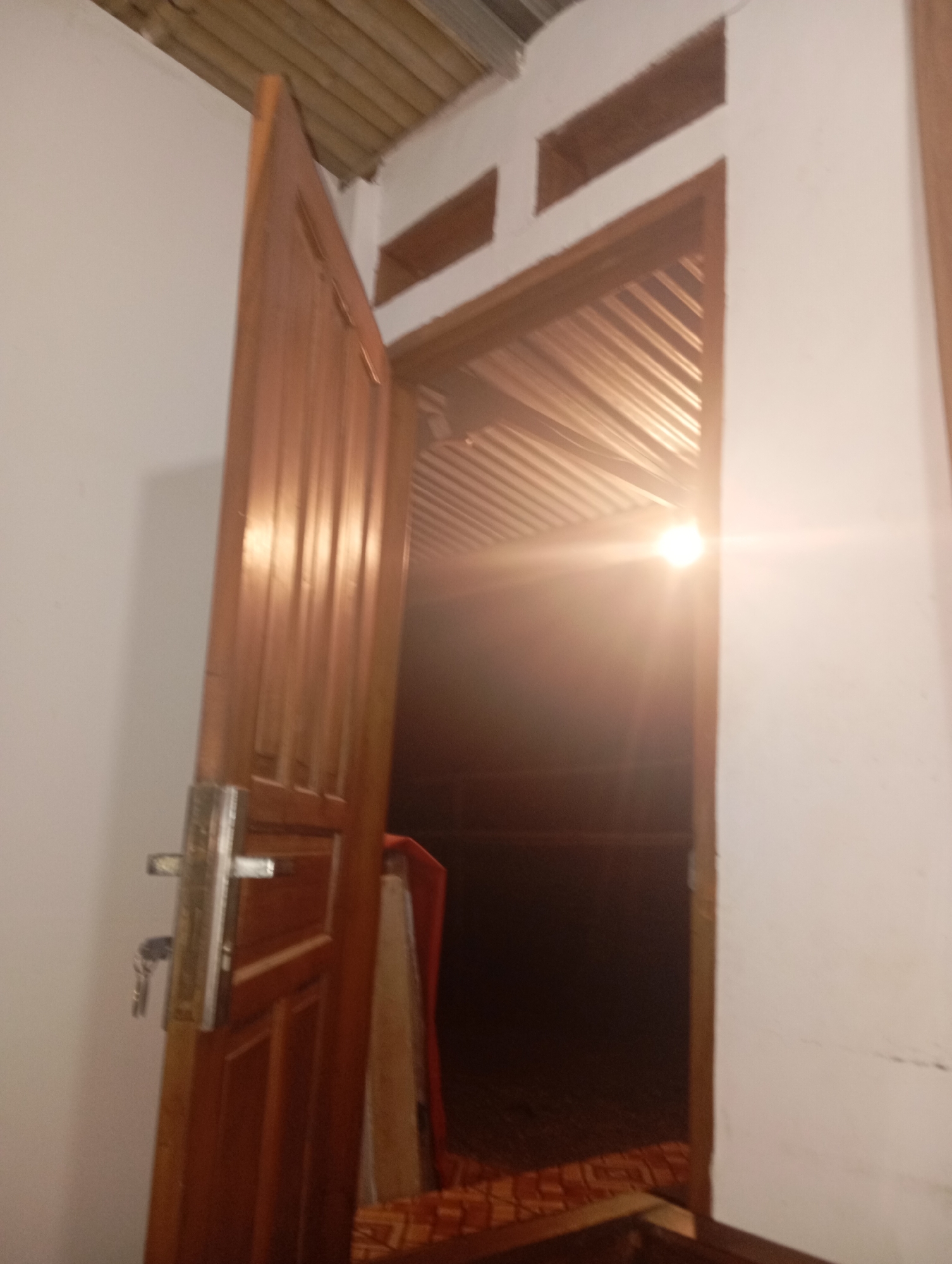 The image depicts a partially open wooden door in what appears to be a building in Kedungbenda, Indonesia. The door, made of polished wood, is ajar, revealing a dimly lit interior. A metal door handle and a set of keys are visible on the left side of the door. Beyond the doorway, the ceiling is covered with corrugated metal sheeting, and a bright light source, possibly a bulb, is emitting a strong glare. The walls of the room appear to be white or off-white. The overall impression is of an interior space, possibly a room or hallway, with a strong light source creating a lens flare effect.