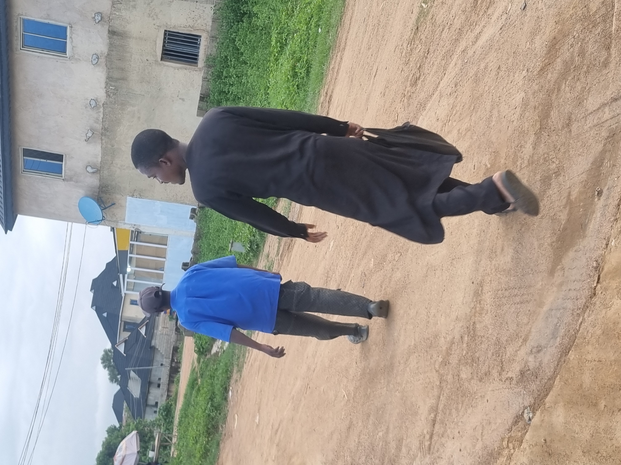 Two men walk away from the camera on a dirt road in Jos, Nigeria. The man in the foreground wears a long, black garment, possibly a traditional robe, and carries a black bag in his right hand. The man behind him wears a blue short-sleeved shirt and dark pants, and a grey baseball cap. The road is flanked by dry grass and vegetation. In the background, several buildings are visible, including a two-story structure with blue-tinted windows and a satellite dish. The sky is overcast, suggesting a cloudy day.