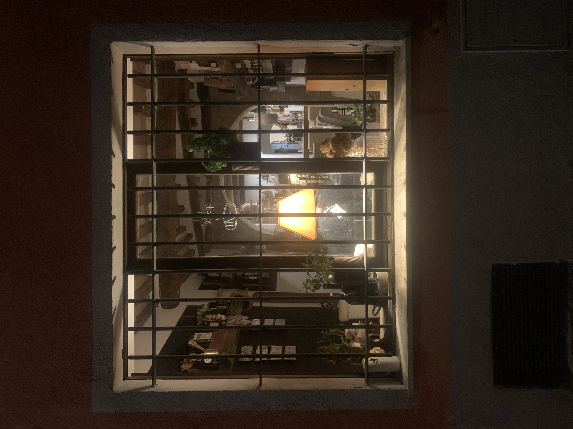The image shows a nighttime view through a barred window into what appears to be a shop or restaurant in San Quirico d'Orcia, Italy. The window has a metal grate, and the interior is illuminated by a warm orange lamp. Shelves are visible behind the grate, stocked with bottles and jars. There are plants and decorative items on display inside. The exterior wall of the building is dark red. Below the window, a dark, rectangular ventilation grate is visible on the lower wall.