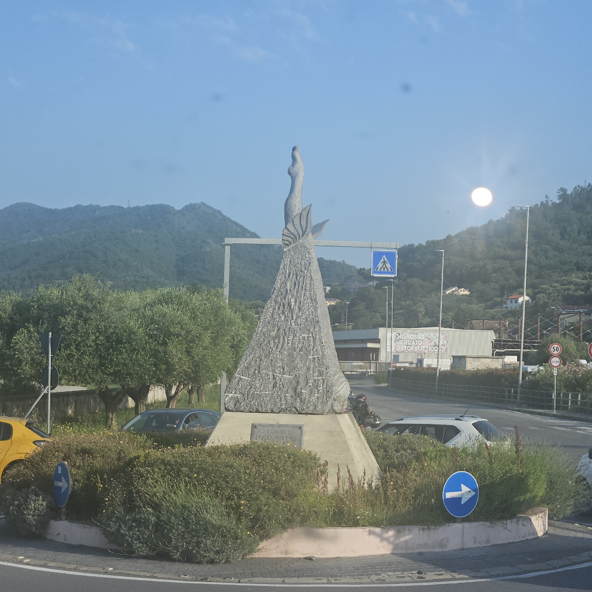The image shows a roundabout in Quiliano, Italy. At the center of the roundabout is a stone monument featuring a slender, abstract human figure atop a textured, conical base. A plaque is visible on the base. The roundabout is encircled by a low, pink-trimmed concrete barrier and filled with green bushes. Cars, including a yellow hatchback, a dark-colored sedan, and a white hatchback, are visible within the roundabout and on the adjacent roads. Street signs, including a pedestrian crossing sign and a blue directional sign, are present. In the background, there are trees, hills, buildings, and a street lamp. A speed limit sign indicating 50 km/h is also visible. The sky is blue with a bright, diffused light source, likely the sun.