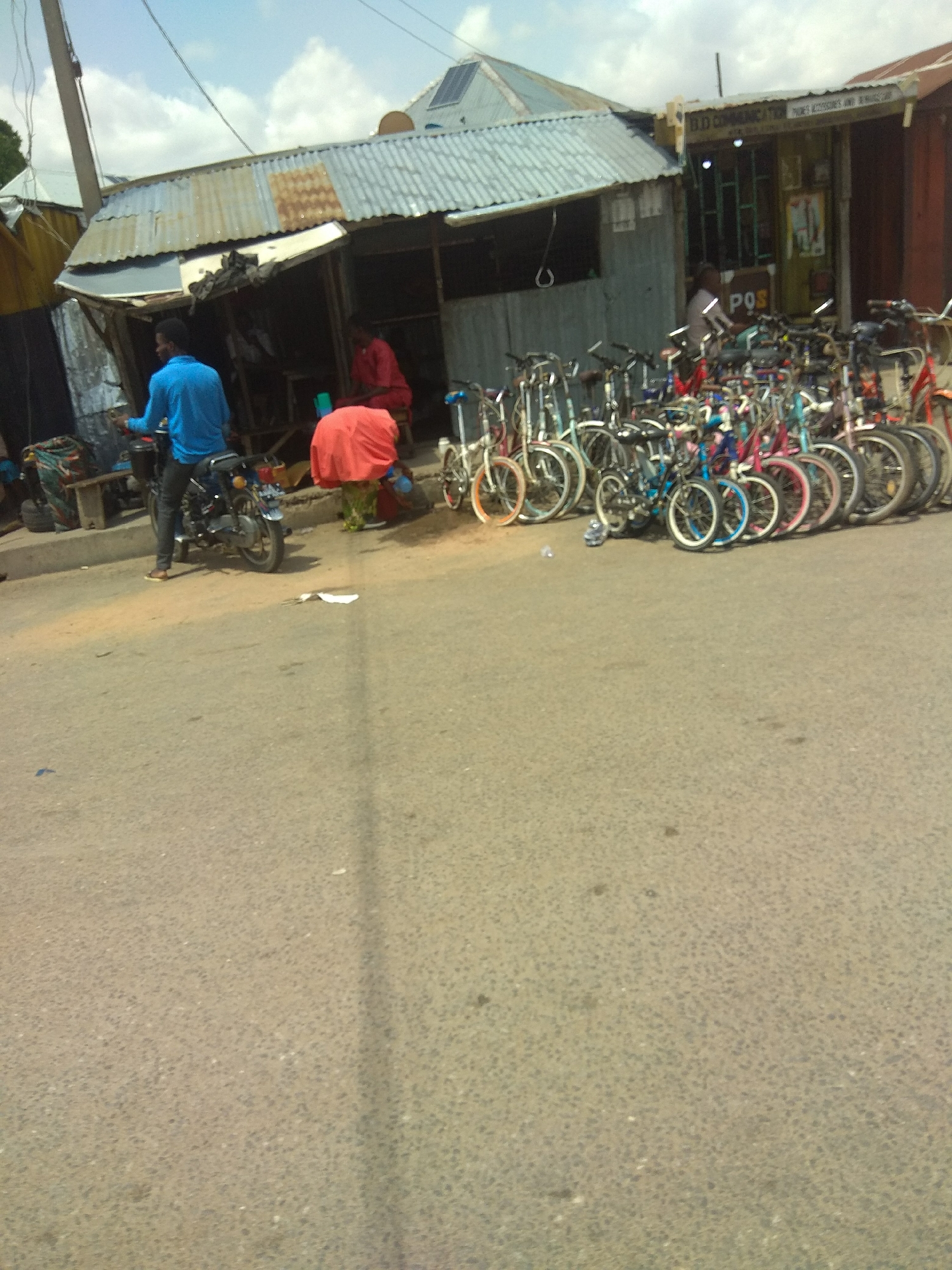 The image shows a street scene in Jalingo, Nigeria, with a focus on a small roadside stall selling bicycles. The stall, constructed from corrugated metal and wood, has a partially open front revealing a dark interior. A person in a blue shirt and dark trousers is seated on a motorcycle in the foreground, facing away from the camera. To the right of the motorcycle, a person in a red garment is seated or kneeling, partially obscured by a large red piece of fabric.
A long row of bicycles, in various colors including white, blue, pink, and red, is parked in front of the stall, suggesting a retail operation. The bicycles appear to be a mix of adult and child sizes, with many featuring basket-like structures. Behind the bicycles, the stall has a sign that reads "POS" prominently displayed, along with other text that is partially obscured. Further into the stall's interior, more items are visible, including what appears to be shelves and possibly other merchandise.
The street itself is paved with asphalt, and there are some debris and discarded papers on the ground. The background shows a cloudy sky and parts of other buildings, including one with a metallic roof and what looks like a solar panel. The overall impression is of a bustling, informal commercial area on a street in Jalingo.