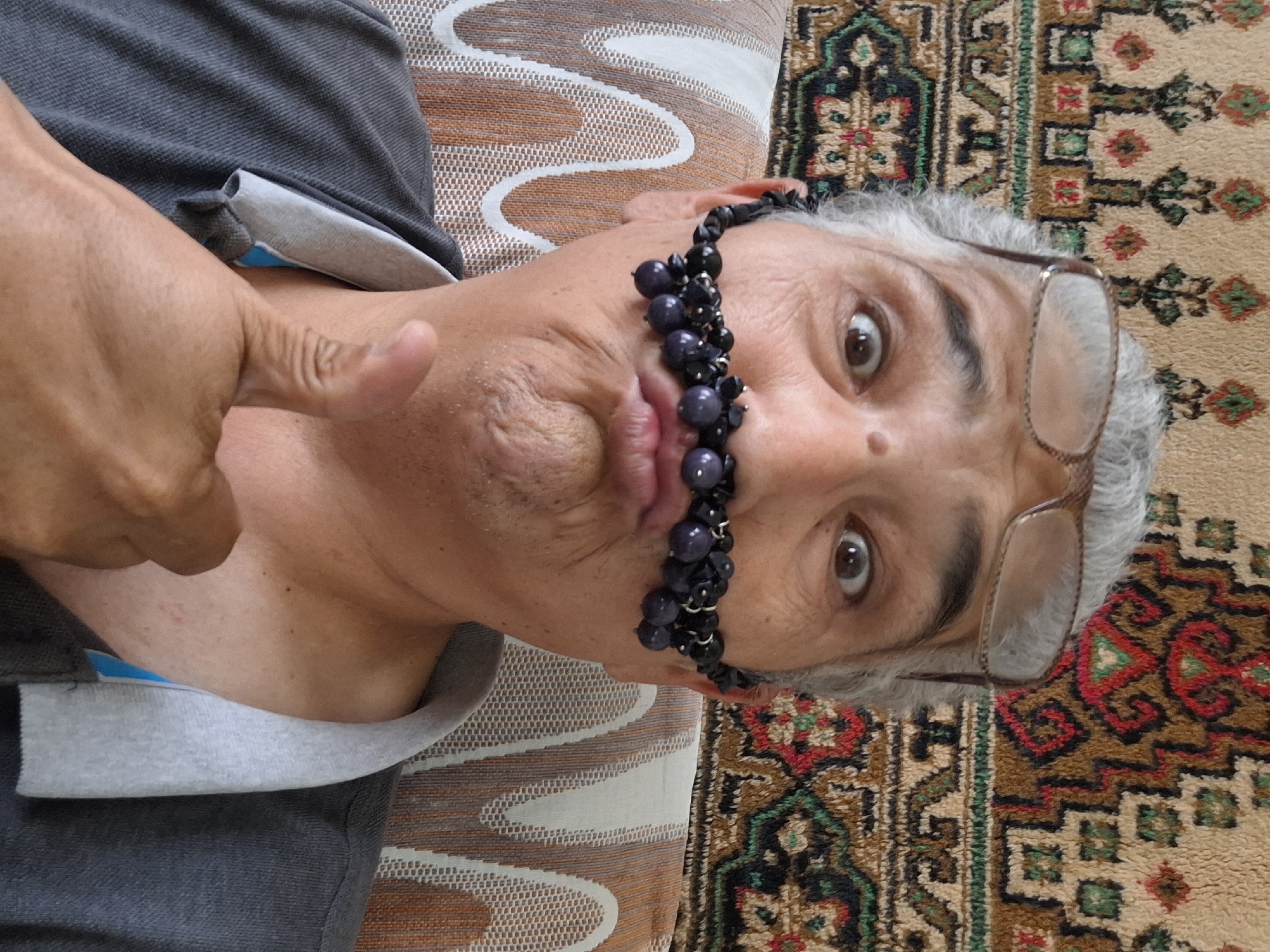 A person with grey hair and glasses is posing for a photo, making a funny face with their lips pursed. They are wearing a grey polo shirt and have a necklace of dark beads draped across their upper lip, resembling a mustache. The background features a tapestry with a geometric pattern and a textured couch. The image appears to be taken indoors. There is no visible indication of the location being Almaty, Kazakhstan.