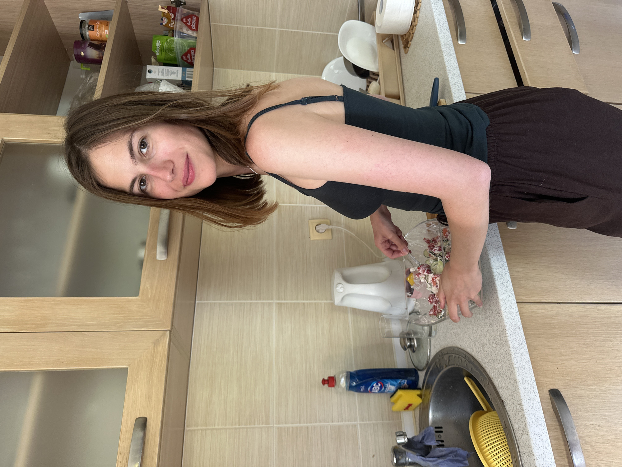 A woman with brown hair is preparing a salad in a kitchen. She is wearing a dark green tank top and brown pants. In front of her, on the kitchen counter, is a glass bowl filled with salad ingredients, and she is using a fork to mix them. To her left, there is a kitchen sink with a blue bottle of dish soap and a yellow sponge. Behind the sink, a white electric kettle is visible. The kitchen features light-colored tiled walls and wooden cabinets. The scene appears to be an indoor residential kitchen.