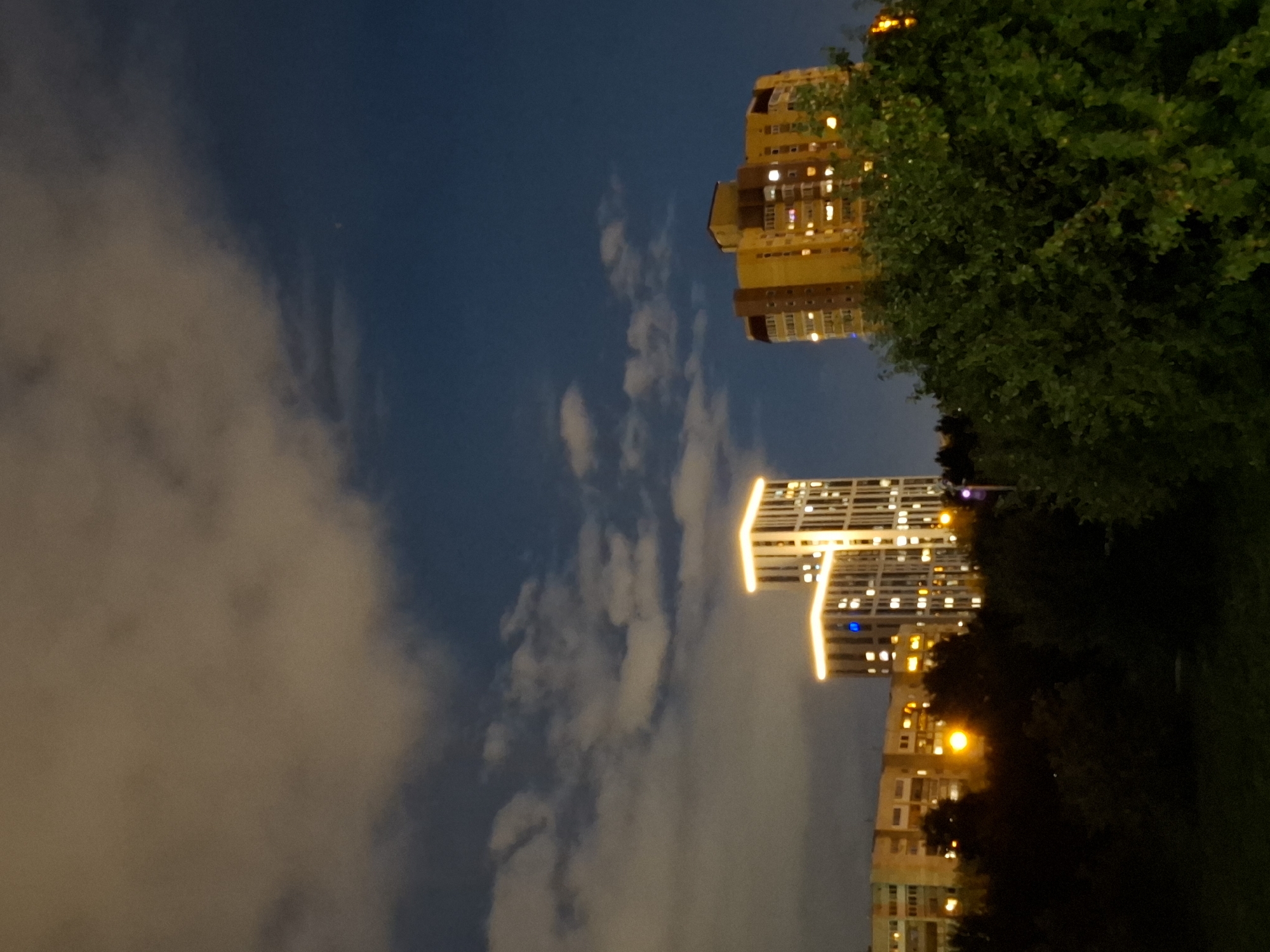 An exterior, low-angle, night-time photograph captures a city skyline featuring several residential buildings and a patch of trees in the foreground. The sky above is a deep blue with scattered clouds illuminated by ambient light. The tallest building, on the left side of the frame, is a modern, white high-rise with prominent vertical lights outlining its structure, and individual windows lit with warm light. To its right, a shorter, light-colored building with visible windows also displays interior lighting. Further to the right, a larger, yellow building stands, with numerous windows lit by interior lights, giving it a warm glow. The foreground is dominated by dark, dense foliage of trees and bushes, with a patch of grass visible at the bottom of the frame. Streetlights emit a warm, orange glow amongst the trees and near the buildings, adding to the illuminated scene.