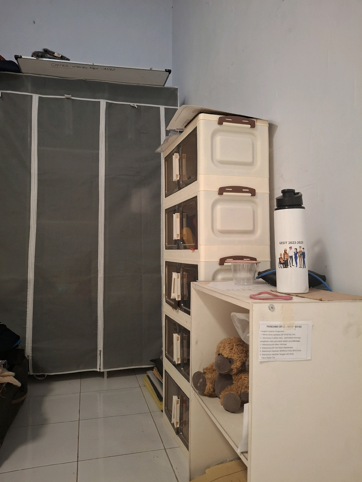 The image depicts an indoor scene, possibly a room in Lumajang, Indonesia, characterized by its mundane and organized clutter. On the left, a tall, gray fabric wardrobe stands, its white zippers and seams prominent against the dark fabric. A whiteboard is propped on top of the wardrobe, with handwritten text that appears to be a list or order.
To the right of the wardrobe, a stack of cream-colored plastic drawers rises, each with a dark brown handle. A transparent plastic cup sits on the top drawer. Further to the right, a white water bottle with a black lid is positioned on a shelf. The bottle features a graphic of stylized figures and the text "GESIT 2023-2025".
Below the drawers and the water bottle, a white shelf unit holds a small, brown, plush toy, likely a teddy bear, on its top shelf. A pink rubber band is visible on the shelf below. A notice with text in Indonesian, including "PANDAWA BPJS", is affixed to the front of this shelf unit. The overall environment suggests a lived-in space, possibly a home or office, with everyday items neatly arranged. The floor is tiled with light-colored squares. The wall behind the objects is a pale blue.