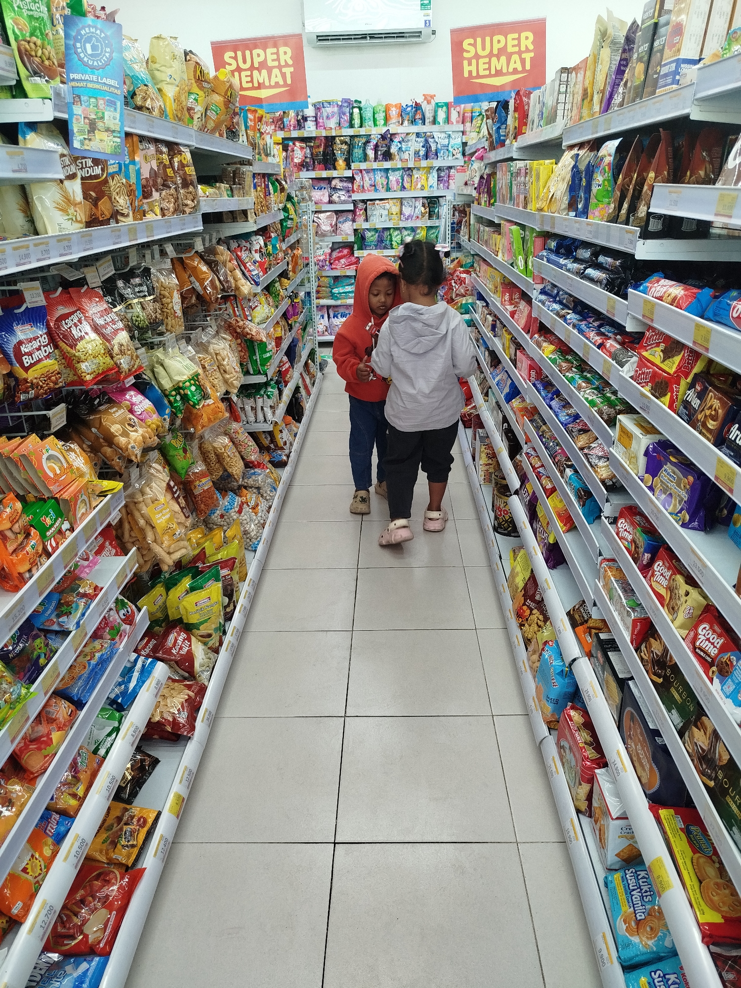 The image depicts the interior of a convenience store in Salamsari, Indonesia, specifically within the aisle designated for snacks. Two young children, a boy and a girl, are the primary subjects. The boy, wearing a red hooded sweatshirt and dark pants, is standing on the left, partially obscured by a shelf. The girl, in a grey hooded sweatshirt and dark pants, is on the right, facing away from the camera and towards the shelves. They appear to be browsing the merchandise.

The shelves on both sides of the aisle are fully stocked with a variety of packaged snacks, including chips, crackers, biscuits, and candies. Colorful packaging is evident throughout. Signs indicating "SUPER HEMAT" (Super Savings) are prominently displayed at the end of the aisle, suggesting a promotional event. The flooring is tiled. The lighting appears to be artificial, typical of an indoor retail environment. The overall scene is one of everyday shopping activity within a well-organized convenience store.