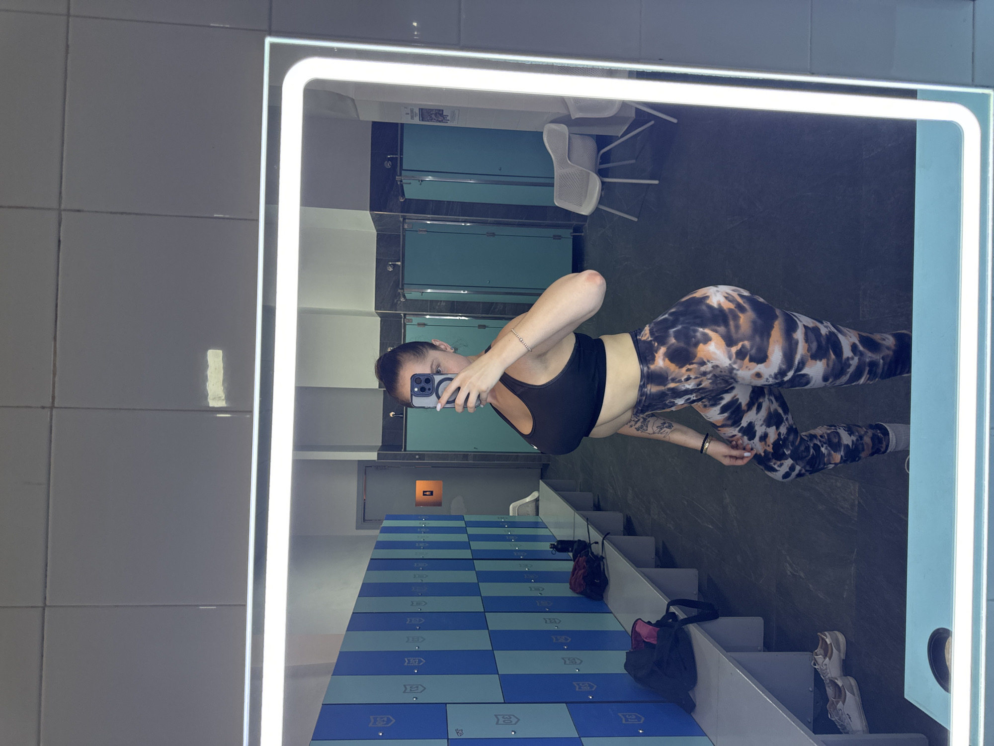 The image shows a woman taking a selfie in a gym locker room. She is wearing a black sports bra and patterned leggings. Her hair is pulled back in a bun. She is holding a smartphone in her right hand and taking the photo in a large, well-lit mirror.
The locker room has light-colored tiled walls and a floor with blue and green checkered lockers. In the reflection of the mirror, we can see gym changing cubicles with teal doors, white chairs, and a white sink. There are also bags and shoes visible on the locker benches. The overall impression is of a clean and modern gym facility.
The image appears to have been taken during the day, as indicated by the bright lighting. There are no obvious weather cues. The woman's expression is neutral, and she seems to be casually documenting her presence at the gym. The setting is clearly a public locker room, likely in a gym or fitness center. The location context of Murcia, Spain, does not provide any specific visual cues within the image itself.
There are some visible numbers on the lockers, such as "3" and "30", likely indicating locker numbers.