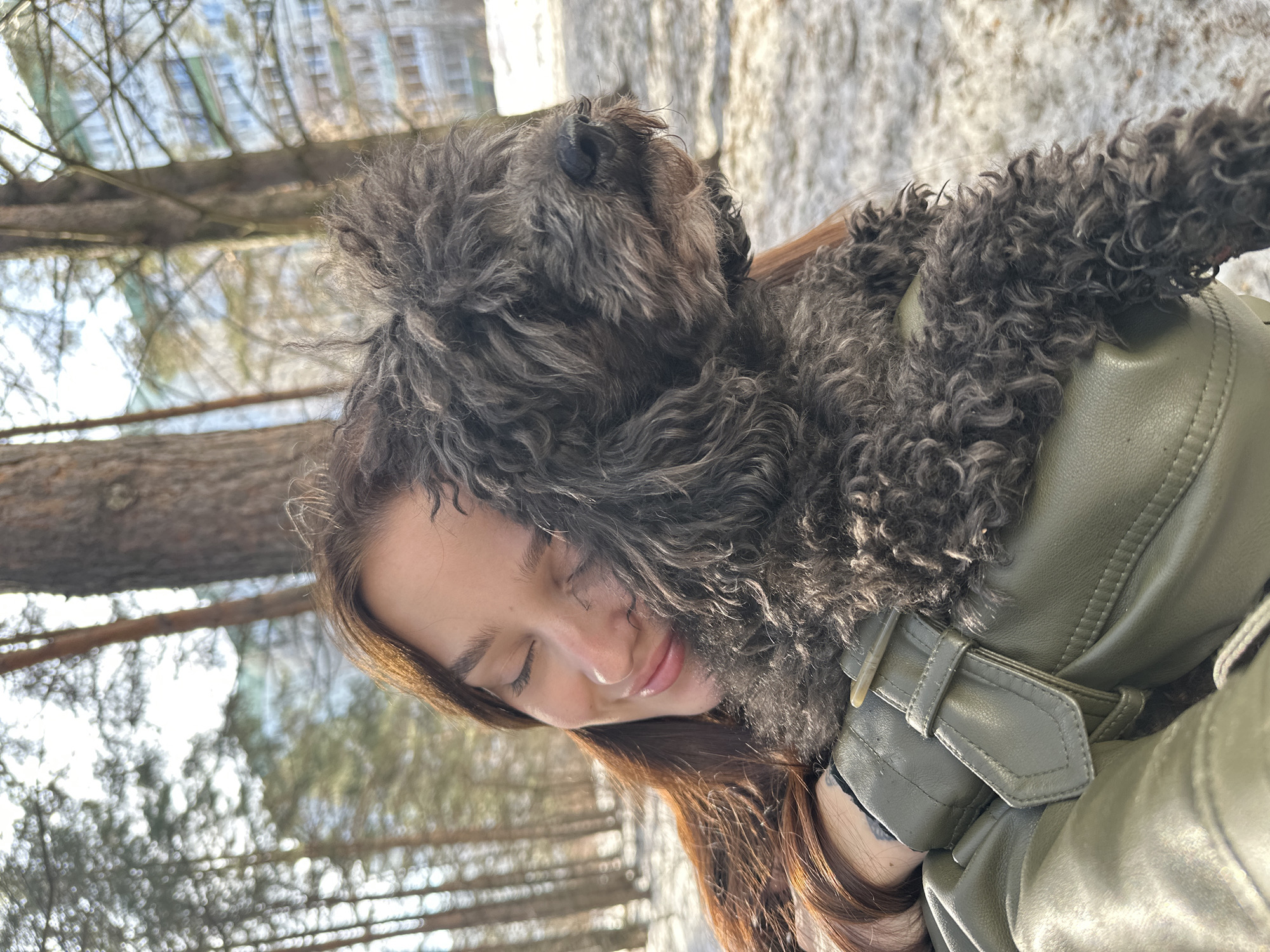 A woman with long brown hair is outdoors in an area with snow on the ground and trees, possibly a park or wooded area in the city of Unknown, Unknown. She is wearing a light green leather jacket and holding a small, dark gray, curly-haired dog. The woman has her eyes closed and appears to be resting her head against the dog. The dog is positioned on the woman's shoulder. The background includes bare trees, snow, and blurred buildings.