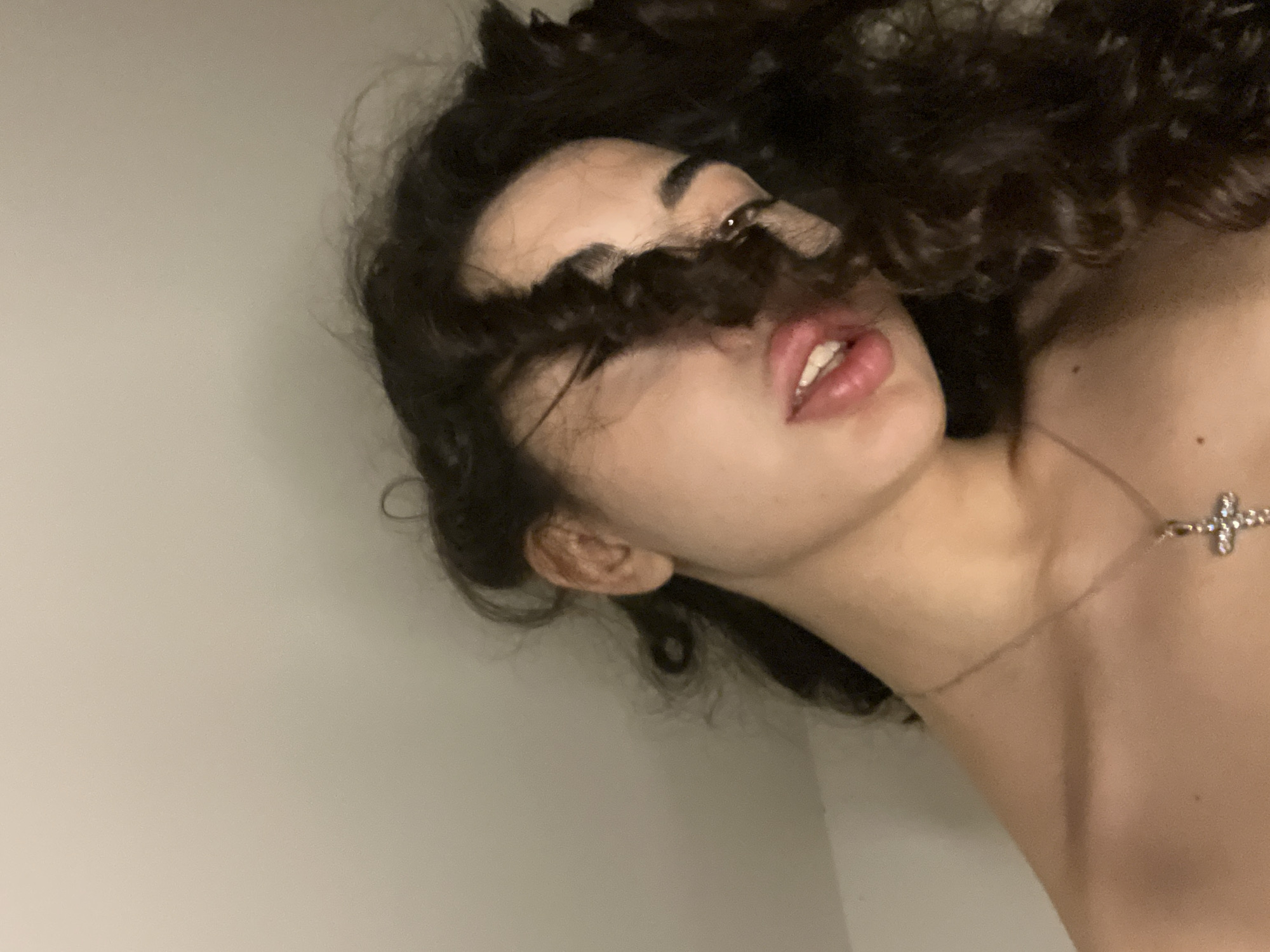 The image depicts a close-up, low-angle shot of a young woman with dark, curly hair. She is looking downwards, with strands of her hair falling across her face and obscuring part of her left eye. Her mouth is slightly open, revealing her teeth. She is wearing a delicate silver necklace with a cross pendant. Her bare shoulders are visible. The background is a plain, light-colored wall, suggesting an indoor setting. There is no identifiable context of Chotěšov, Czechia, within the image.