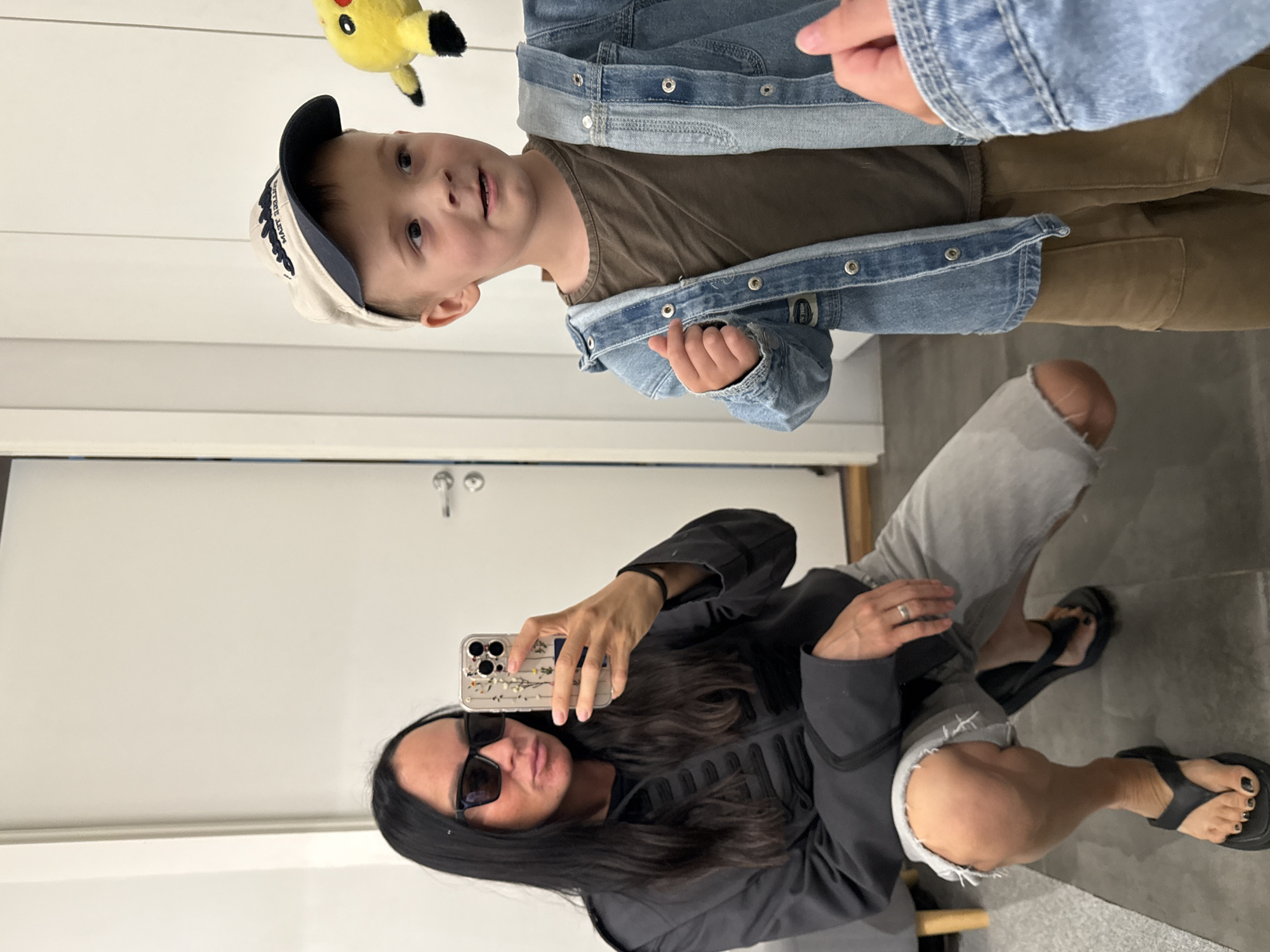 The image depicts a young boy wearing a baseball cap, a brown t-shirt, and a denim jacket, holding a yellow Pikachu plush toy. He is standing in front of a white door, possibly in an apartment. To the left, a woman with long dark hair and sunglasses is taking a selfie with her phone. She is wearing a dark jacket and light-colored ripped jeans, and black flip-flops. The setting appears to be indoors, with a closet door visible in the background and part of a sofa. There is no indication of the city of Chaikovsky, Russia, or any other specific urban location.