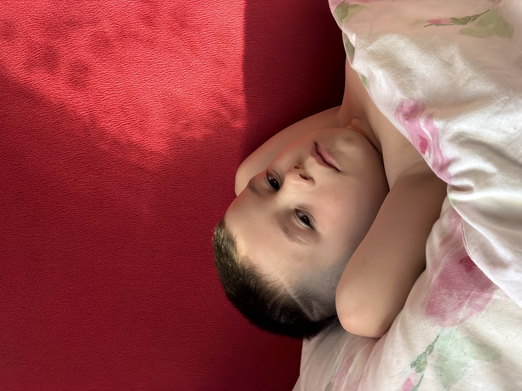 The image displays a young male child, estimated to be between 4 and 7 years old, lying on his back in an indoor setting. The specific location within the city of Chaikovsky, Russia, cannot be determined from the provided visual information. The child has short, dark hair styled with a faded undercut on the sides, featuring a distinct shaved line above the left temple. He rests his head on a white pillow while both hands are positioned behind his head. His eyes are open, and he is looking directly towards the viewer with a neutral facial expression. He is partially covered by a white blanket or duvet patterned with pink roses and green leaves. The background behind the child's head is a solid, deep red, textured surface, appearing to be a headboard or an adjacent wall. A strong, diagonal beam of light, consistent with sunlight, illuminates the upper right portion of this red background, creating a clear line of shadow below it. No other individuals or specific identifying objects are visible within the frame.