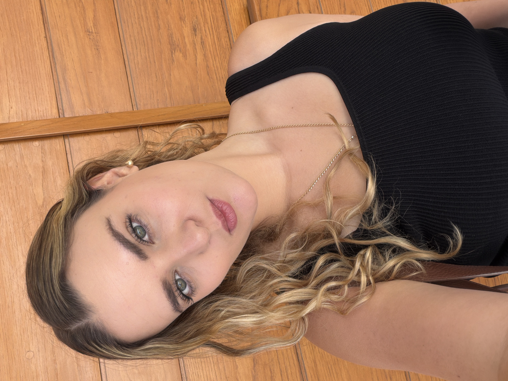 A medium close-up portrait shows a woman posing indoors in San Bernardino Tlaxcalancingo, Mexico. She is lying down, with her head tilted to her right, looking directly at the viewer. Her light-brown to blonde hair, styled in loose waves and curls, cascades around her head and shoulders. She has striking green eyes with visible mascara on her lashes, and her eyebrows are well-defined. Her lips are slightly parted, revealing a subtle pink lip color. She is wearing a ribbed black top or dress with a high neckline. A delicate gold chain necklace is visible around her neck, and a small gold stud earring can be seen on her right earlobe. The background consists of warm-toned wooden planks, possibly a floor or wall, with a distinct horizontal wooden strip running across the frame. The lighting appears soft and even, suggesting an indoor setting with diffused light. No text is visible in the image.