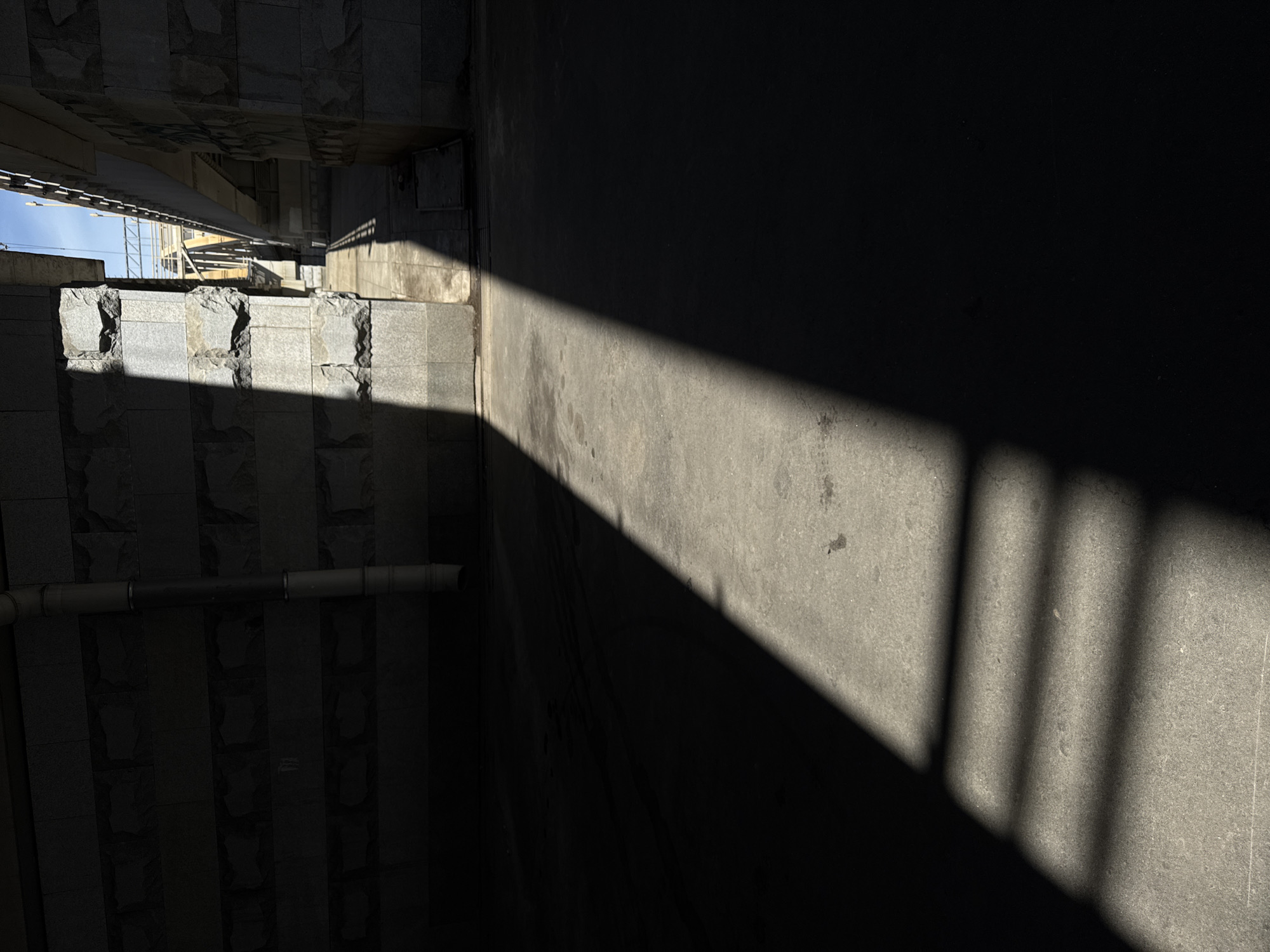 The image captures an urban scene in Moscow, Russia, characterized by strong contrasts of light and shadow. The primary subjects are architectural elements, specifically a textured stone wall on the left and a concrete surface on the right, bisected by a sharp, diagonal beam of sunlight. On the left, a layered stone structure is visible, appearing to be part of a building or an underpass, with its rough-hewn surfaces catching the light in some areas and remaining in deep shadow in others. A horizontal pipe runs across this section.
The right side of the image is dominated by a large, dark shadow cast by an unseen object, creating a stark division with the sunlit concrete. Within this sunlit area, the shadow of a railing or bars is cast, creating a repeating pattern of vertical lines. The overall impression is one of an outdoor, possibly public space, with elements of construction or infrastructure.
The lighting suggests a time of day when the sun is relatively low, creating long, defined shadows. This could be morning or late afternoon. The weather appears clear and sunny, indicated by the bright, direct light. There are no people visible in the frame. The textures of the stone and concrete are prominent, adding to the tactile quality of the scene. The emotional tone is stark and geometric, emphasizing form and light rather than human presence. No text is visible in the image.