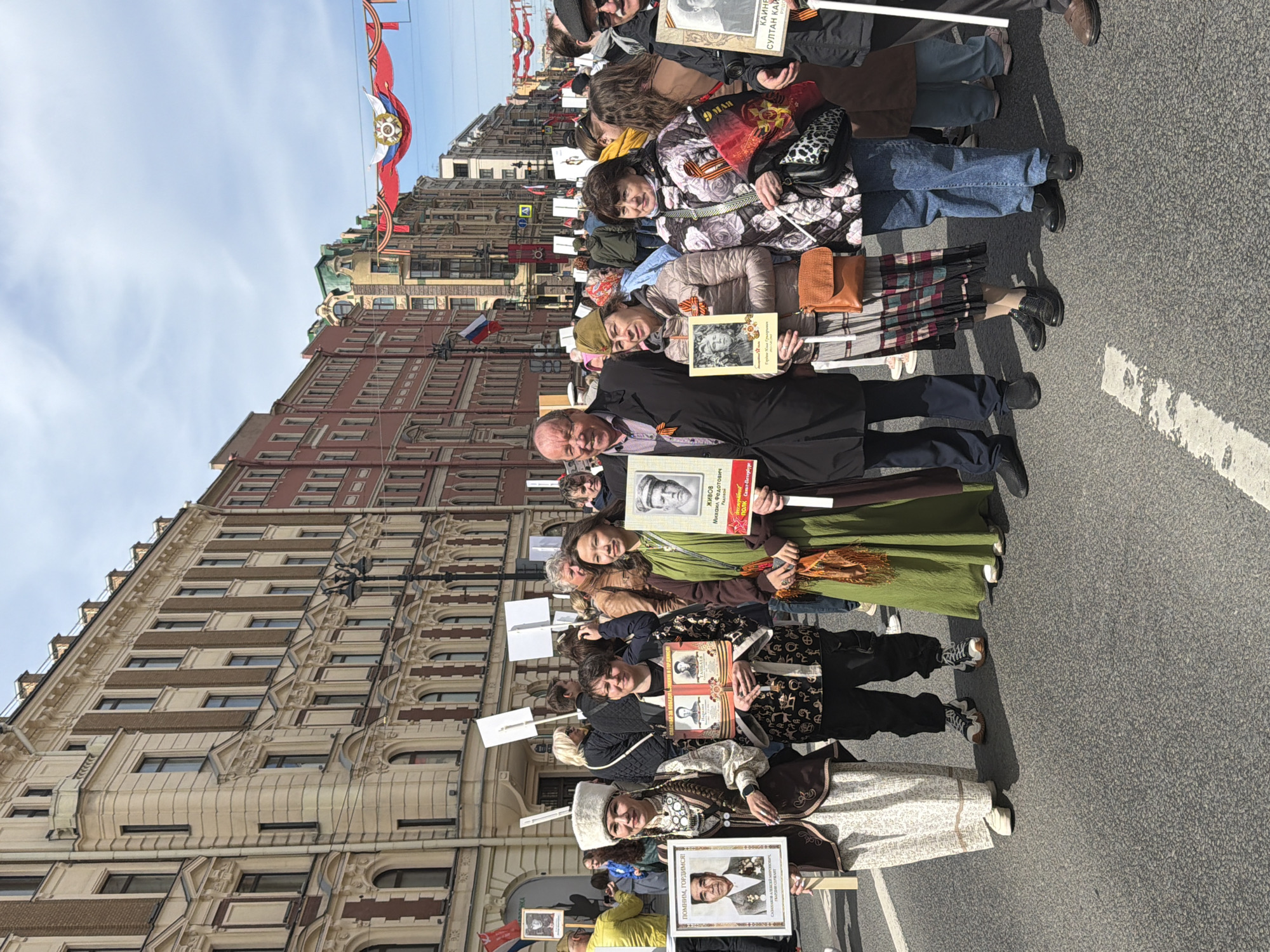 A procession of people is holding up images in frames of military personnel, with a series of Russian flags displayed on overhead wires. The crowd is assembled on a street in Saint Petersburg, Russia, and a sign reads "Remember, be proud!" in Russian. Several other building frontages are visible in the background. The people are standing on the pavement.