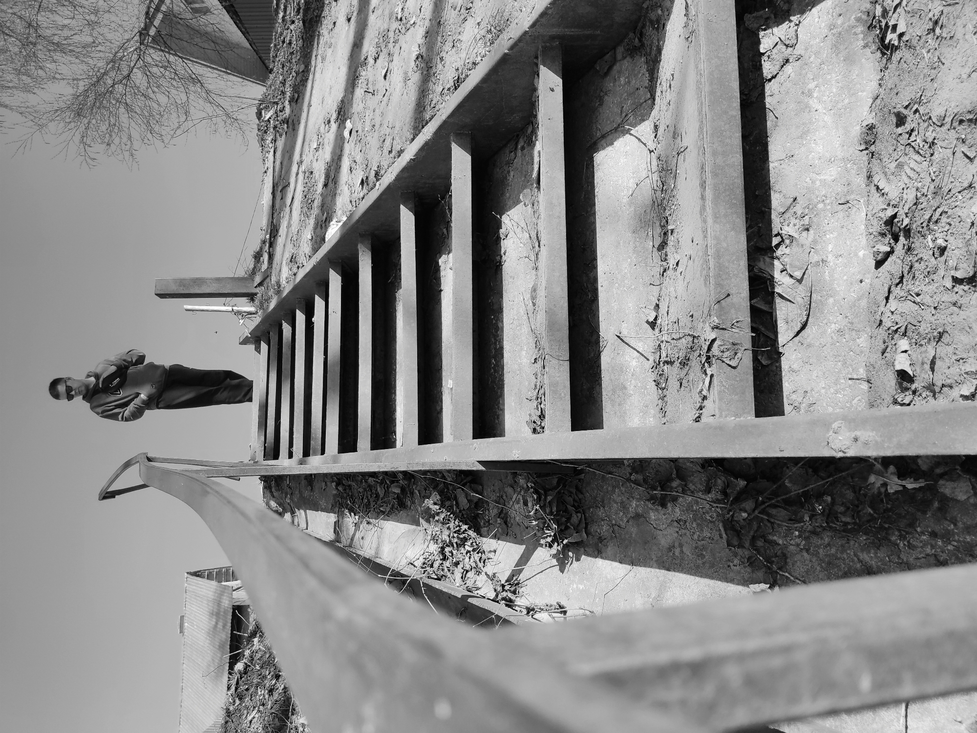 The image, in black and white, presents an unusual, tilted perspective, potentially from a low angle. It features a metal railing and a ladder-like structure attached to a rough, possibly concrete wall covered in dirt and debris. A man is oriented upside down against a clear sky in the upper left portion of the frame. He appears to be wearing sunglasses, a hooded jacket, and trousers, with one hand seemingly holding or adjusting something. Some bare tree branches can be seen in the very top left corner. Parts of buildings are seen in the background. Considering the provided information, it can be assumed this scene is located in Chaikovsky, Russia.
