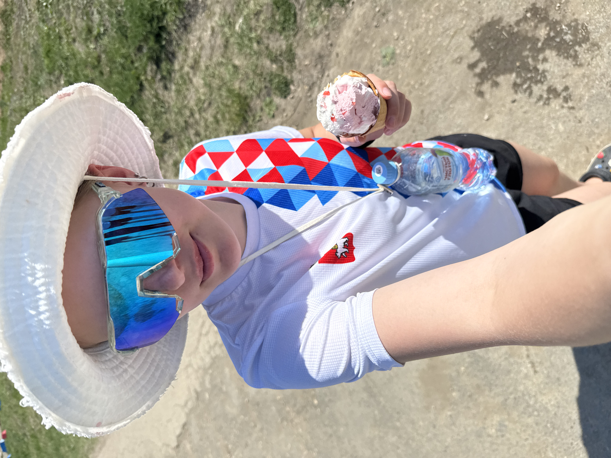A person is sitting outdoors in Khvalynsk, Russia, holding an ice cream cone. They wear a white sun hat, blue reflective sunglasses, a white shirt with the city of Khvalynsk's coat of arms, a red and blue patterned shoulder wrap, and black shorts. A water bottle is placed near their side. The ground appears to be concrete or paved.