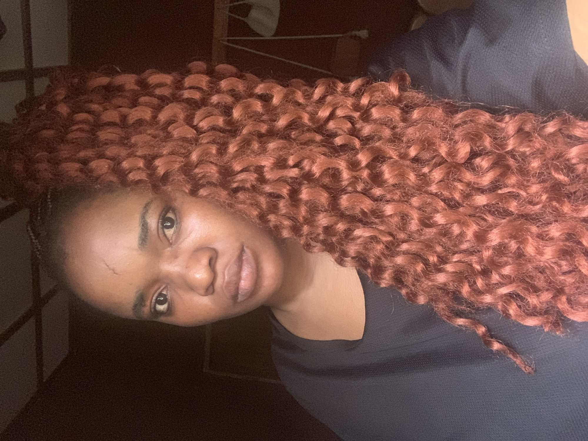 A portrait-style image captures a Black woman from the chest up, oriented sideways. She has warm, reddish-brown hair styled in long, voluminous curls, possibly extensions or braids, that cascade down one side of the frame. Her hair is a prominent feature, with distinct, well-defined coils.
She is wearing a simple black top. Her face is turned towards the viewer, with her gaze directed at the camera. Her eyes are dark, and her lips appear full. There is a visible mark or scar on her forehead. The lighting is dim, suggesting an indoor setting with low ambient light, potentially evening or night. The background is dark and indistinct, with faint structural lines visible in the upper left corner, possibly part of furniture or a window frame. The overall atmosphere is intimate and softly lit. The woman's expression is neutral and direct. The image is set in Jos, Nigeria. No visible text is present.