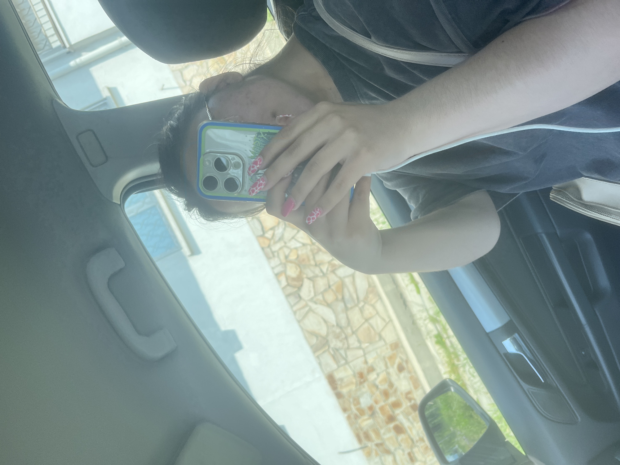 The image, rotated 90 degrees counter-clockwise, depicts a young woman seated inside a car, holding a smartphone and seemingly in the act of taking a selfie. Her head is tilted, and her face is largely obscured by the back of the phone, though parts of her dark hair, forehead, and cheek are visible. She is wearing a dark gray or black top with a lighter strap across her shoulder.
She holds an iPhone-like smartphone with a clear case that features a vibrant design of pink flowers or strawberries and green leaves. The phone's multi-lens camera system is prominent. Her fingernails are long and manicured, painted in a striking pink color with distinctive white cow-print or spotted patterns. The partial word "NORMAN" or similar is faintly visible on the phone case near the camera bump. A white charging cable extends across her arm, and a light-colored purse or bag is visible beside her.
The car's interior is bright, indicating daylight. Visible car elements include the light-colored ceiling with a grab handle, part of the A-pillar, and a dark door panel featuring a chrome handle. Through the car windows and reflections, an outdoor scene is visible, including a light-colored building, a wall constructed from irregularly shaped, light brown and beige stones, and some green foliage. The bright light suggests it is daytime, likely midday or afternoon, under clear weather conditions. The setting appears to be an urban or suburban area in Varna, Bulgaria.