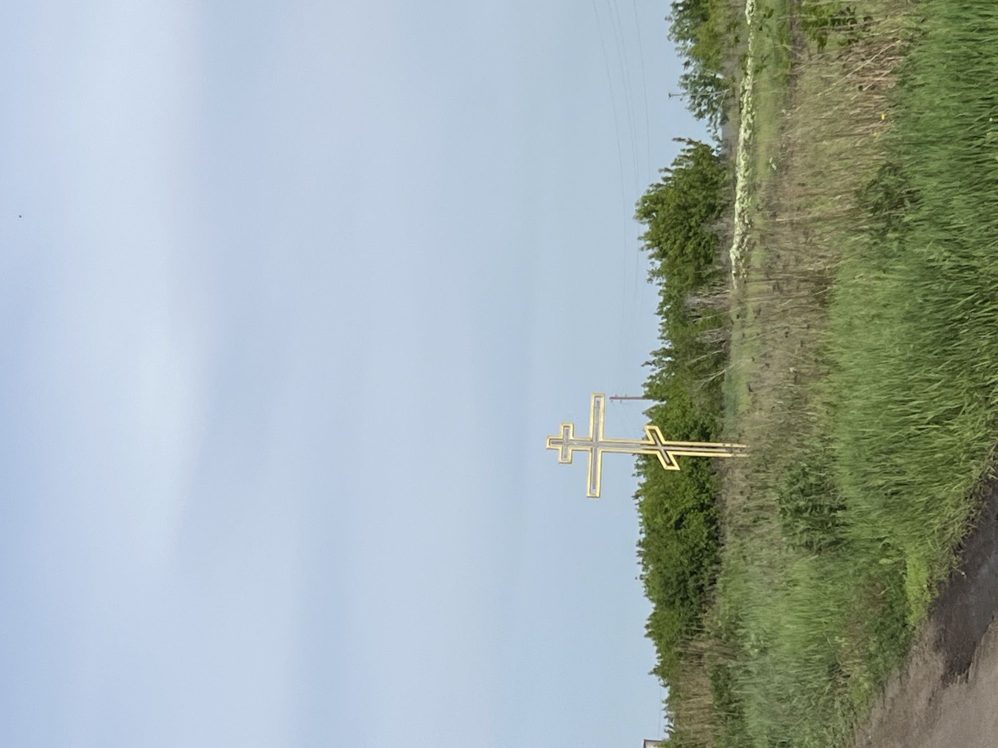 A vertical shot shows a gold Eastern Orthodox cross set in overgrown grasses along a road outside Vysoke, Ukraine. The background features a light blue sky and utility lines at the top right corner of the frame.