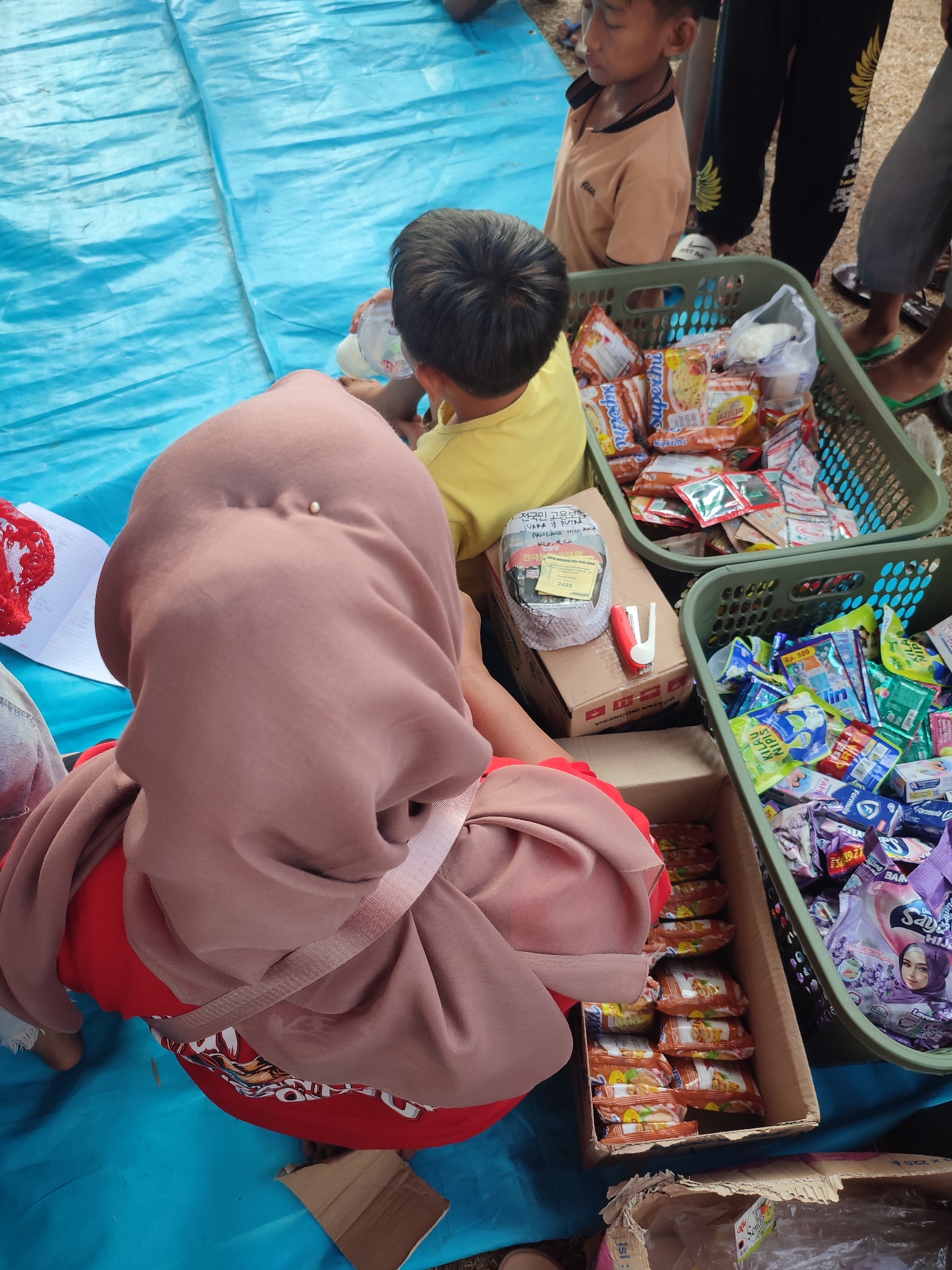 In Nusa Jaya, Indonesia, a group of people, including children and an adult wearing a hijab, are gathered around a blue tarp. The adult, facing away from the camera, is wearing a red shirt and a brown hijab. A child in a yellow shirt is seated nearby, and another child in an orange shirt stands to the left, looking down.
Two green plastic baskets overflow with various packaged food items and toiletries. One basket contains individual sachets of what appear to be food seasonings and instant noodles. The other basket is filled with small packages of candy, soap, and laundry detergent. A cardboard box sits between the two baskets, with a red stapler and some papers on top, one of which has handwritten text in Korean. Another cardboard box is visible in the foreground, partially open and containing plastic bags. The scene suggests a distribution or sharing of goods, possibly a community event or aid distribution in the city.